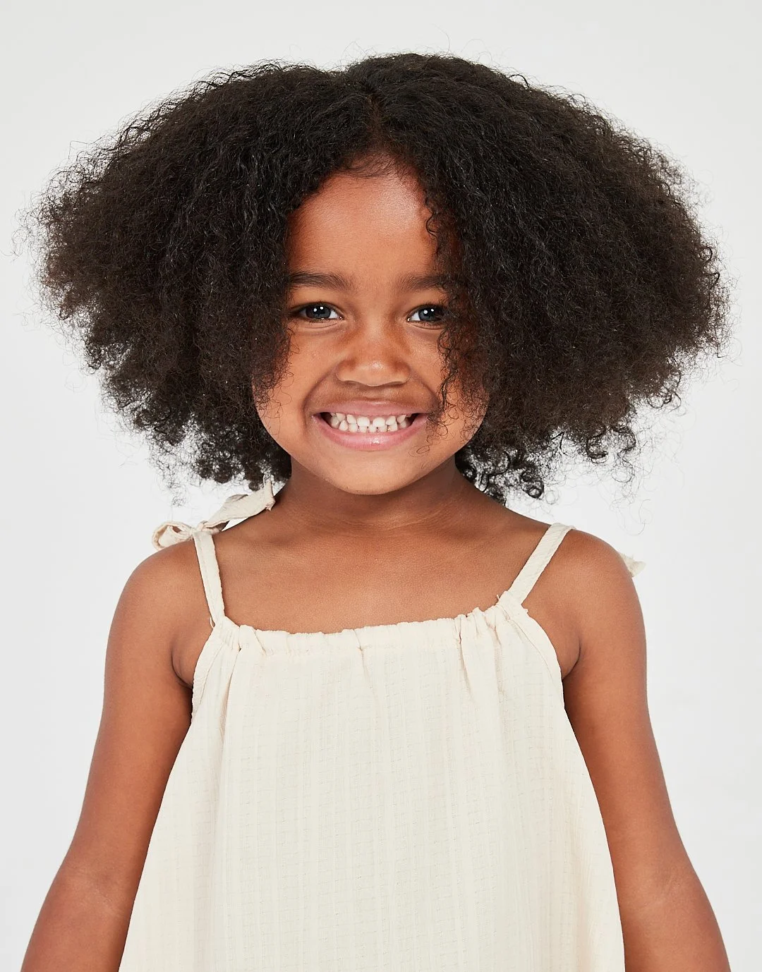 A young girl with curly black hair smiling and showing her teeth, wearing a beige dress with spaghetti straps.