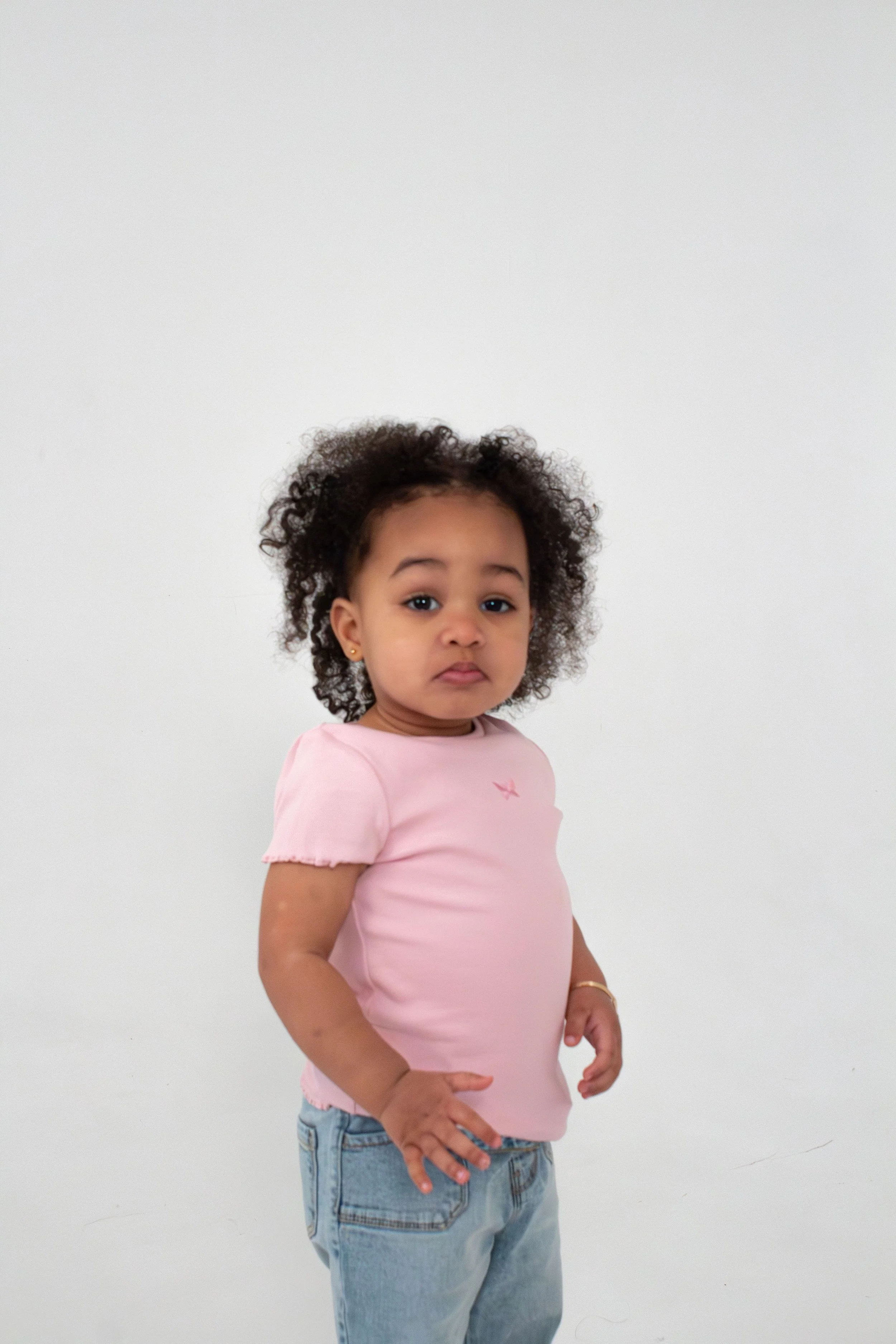 Young girl with curly hair wearing a pink shirt and light blue jeans standing against a white background.