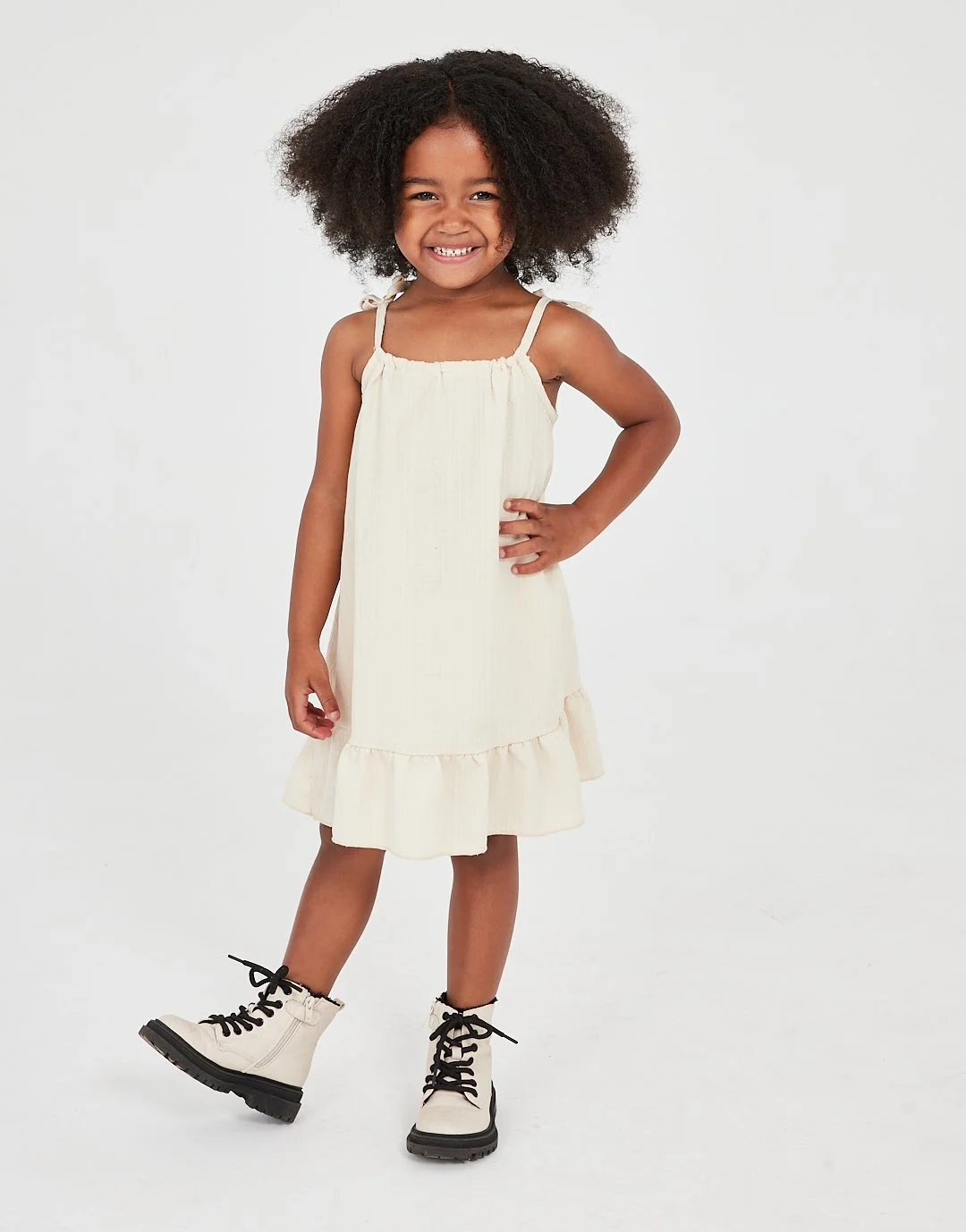 Young girl smiling, wearing a cream-colored dress with ruffle hem and black combat boots with laces, standing with one hand on her hip against a white background. Child model for Socityuk.