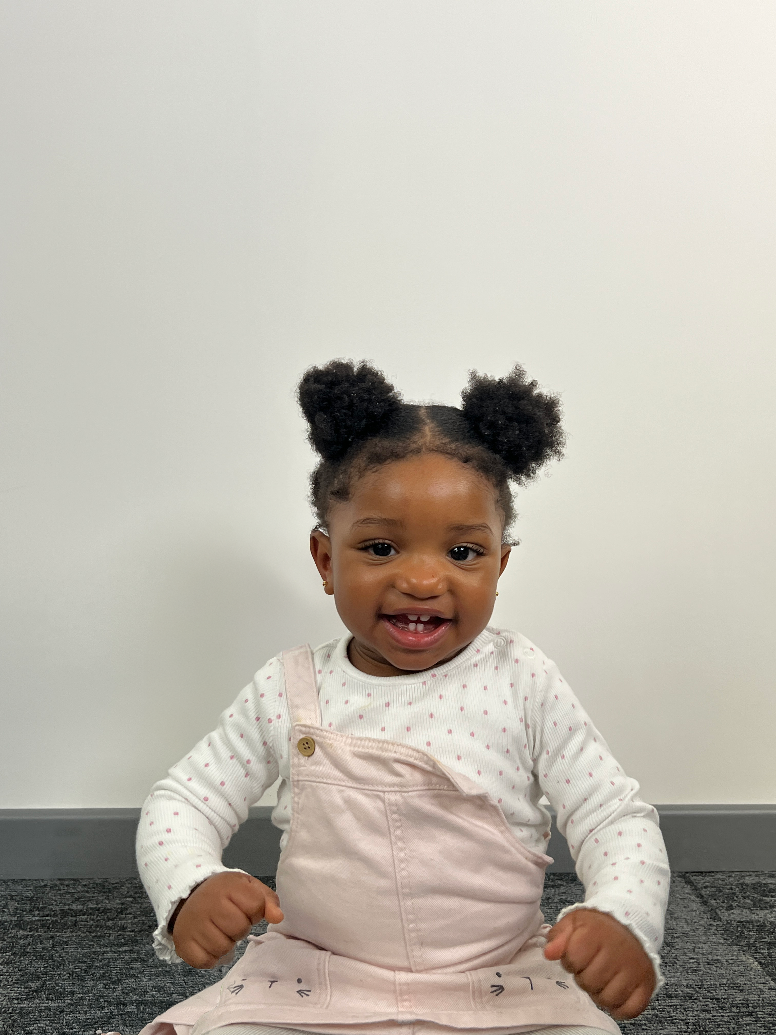 Smiling young girl with dark curly hair in puffy buns, wearing a white shirt with pink polka dots and pink overalls, sitting on a gray carpet against a plain white wall.