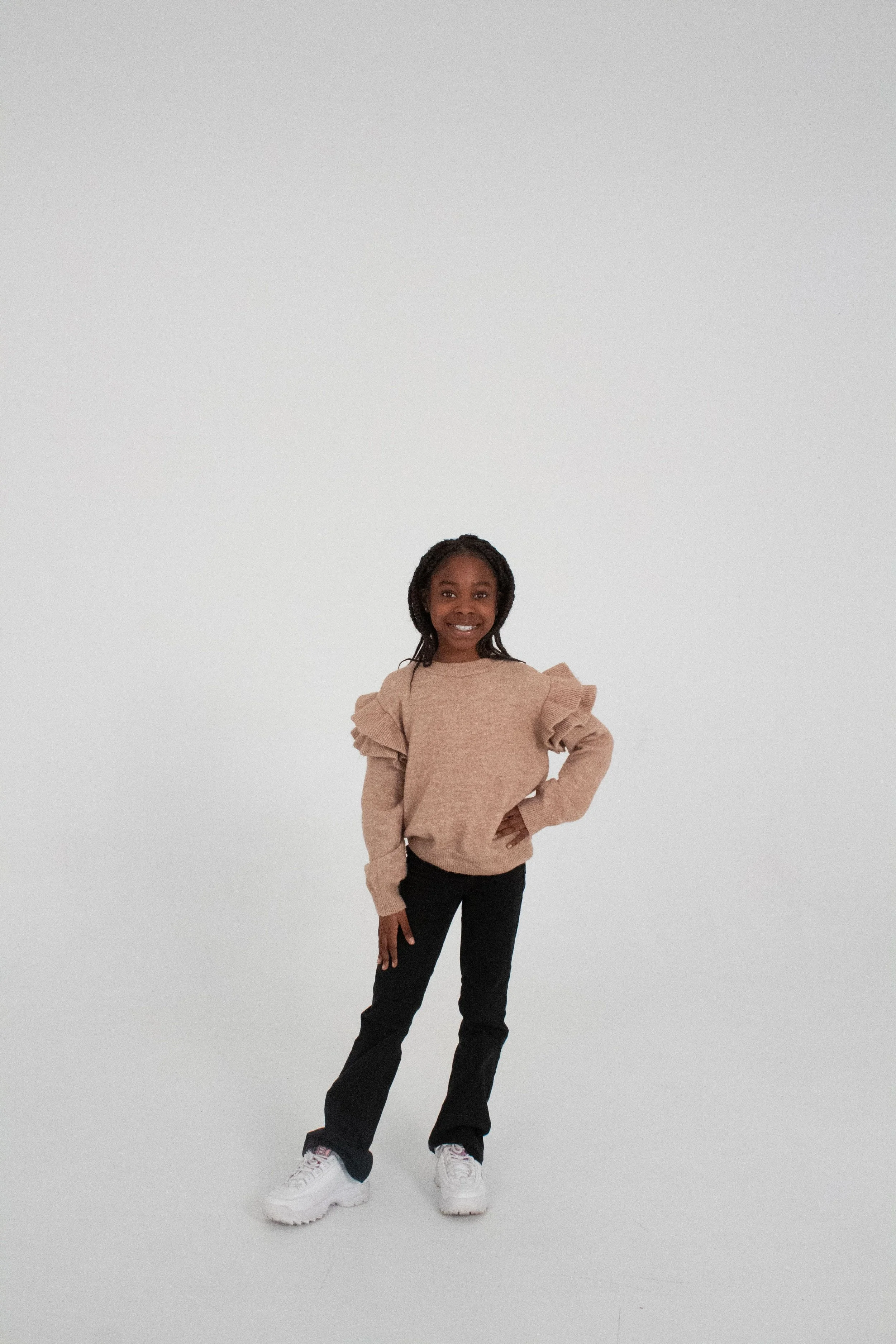 A young girl standing confidently with one hand on her hip, wearing a beige sweater with puffed sleeves, black pants, and white sneakers, against a plain white background.