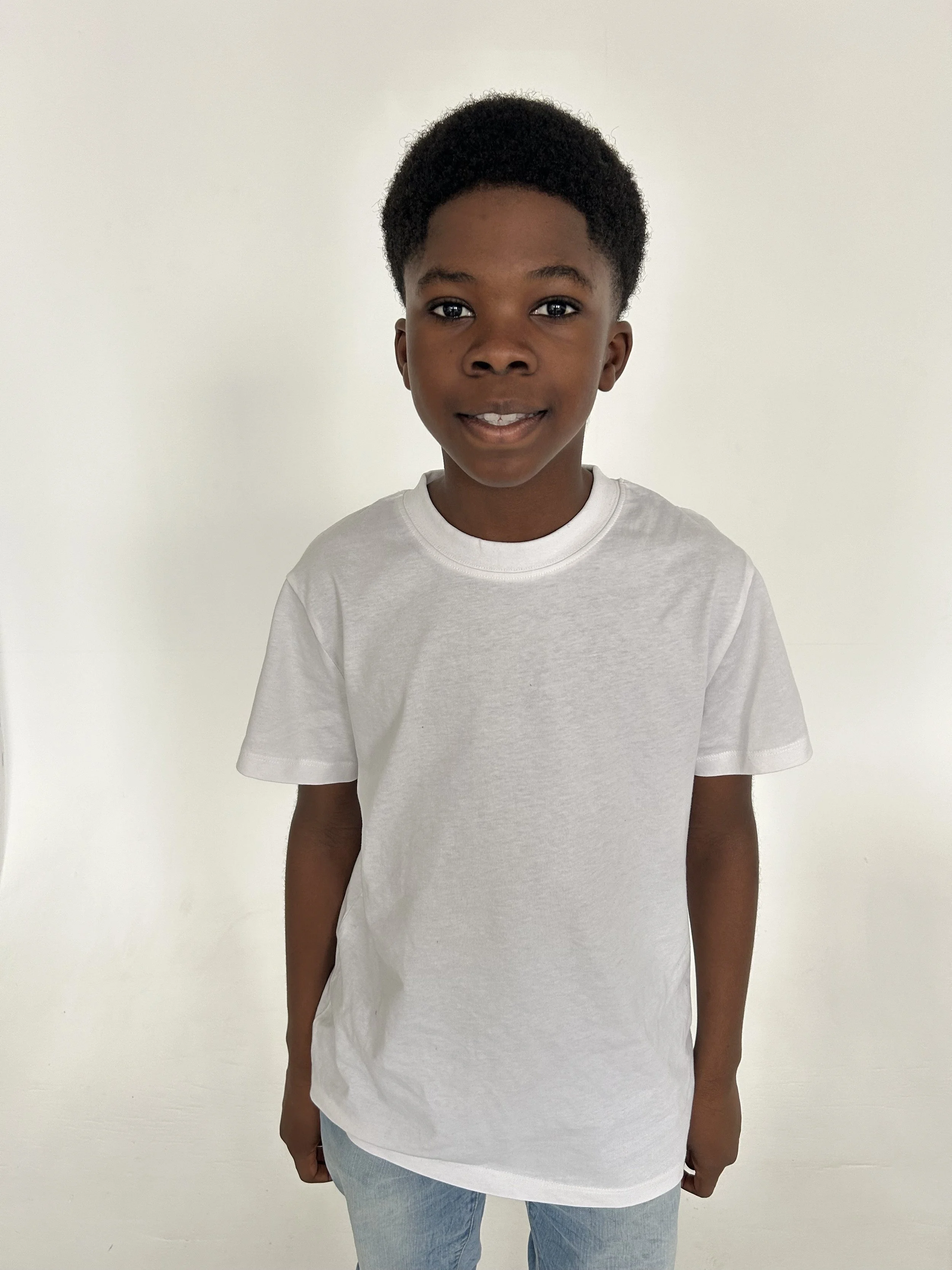 A young boy with dark skin, curly black hair, wearing a plain white t-shirt and light blue jeans, standing in front of a plain white background and smiling slightly. Model for leading Manchester and London based child model agency Socityuk.