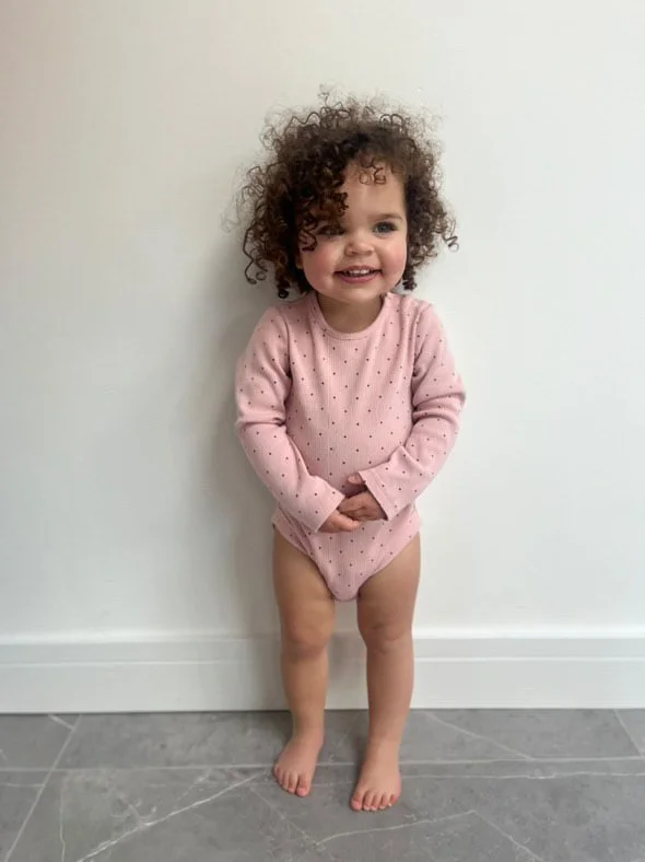 A young girl with curly hair smiling and wearing a pink long-sleeve shirt with polka dots, standing barefoot against a white wall. Modelling for leading child model and talent agency SocityUK based in London and Manchester.