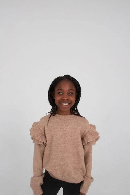 Smiling young girl with braided hair, wearing a beige sweater with ruffled sleeves, standing against a plain white background.