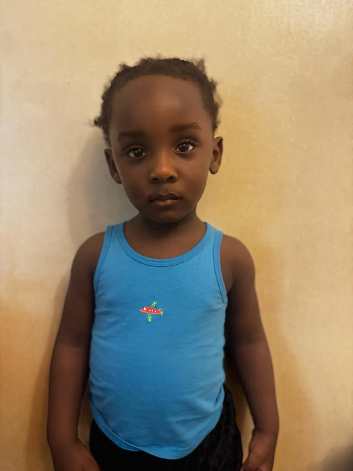 A young boy with short hair and dark skin wearing a blue sleeveless shirt with a small aircraft graphic, standing against a plain beige wall. Modelling for leading child model and talent agency SocityUK based in London and Manchester.