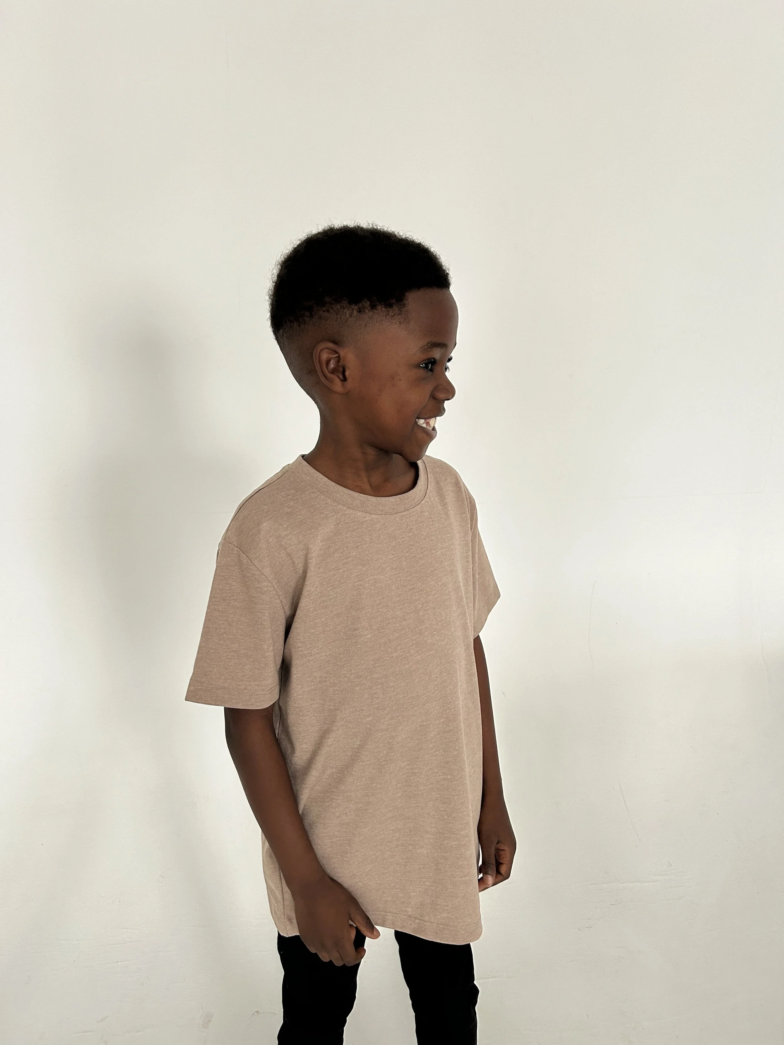 Young boy with short curly hair smiling and standing sideways against a plain white wall, wearing a beige T-shirt and black pants. Model for leading Manchester and London based child model agency Socityuk.