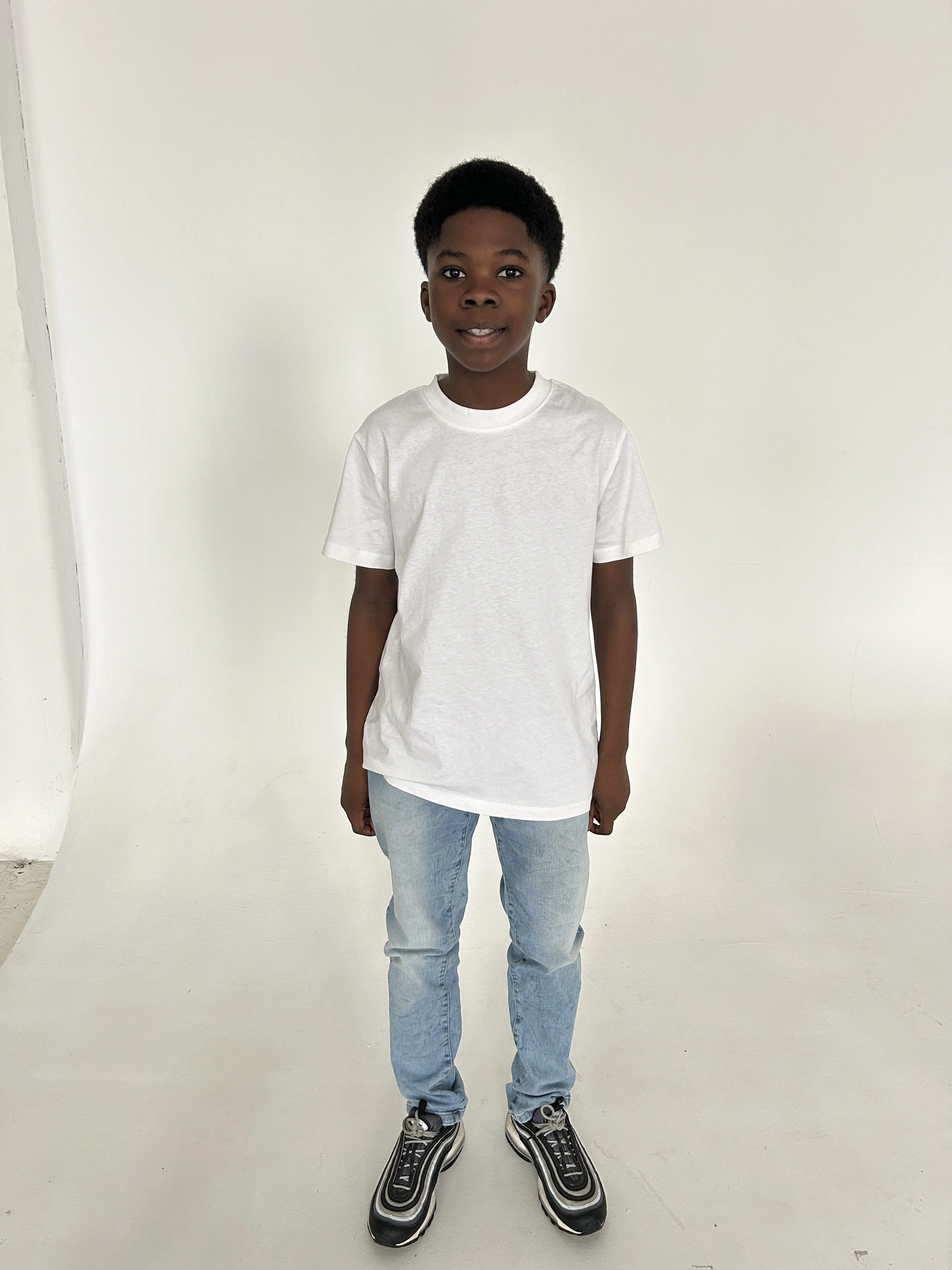 A young boy with dark skin and short curly hair standing in front of a plain white background, wearing a white t-shirt, light blue jeans, and black sneakers.