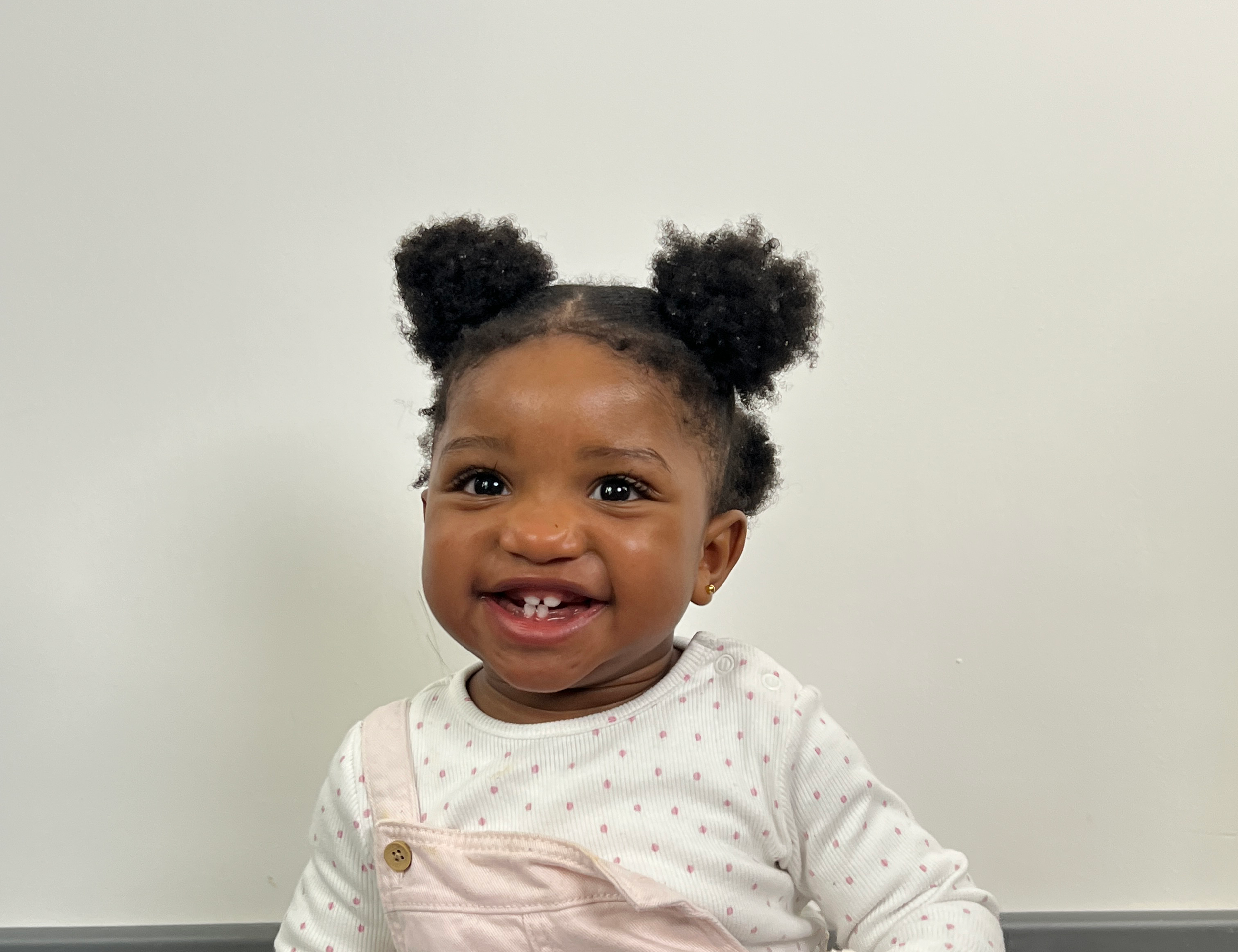 A smiling young girl with dark hair styled in two puffy buns, wearing a white polka dot shirt and light pink overalls, standing against a plain white wall.
