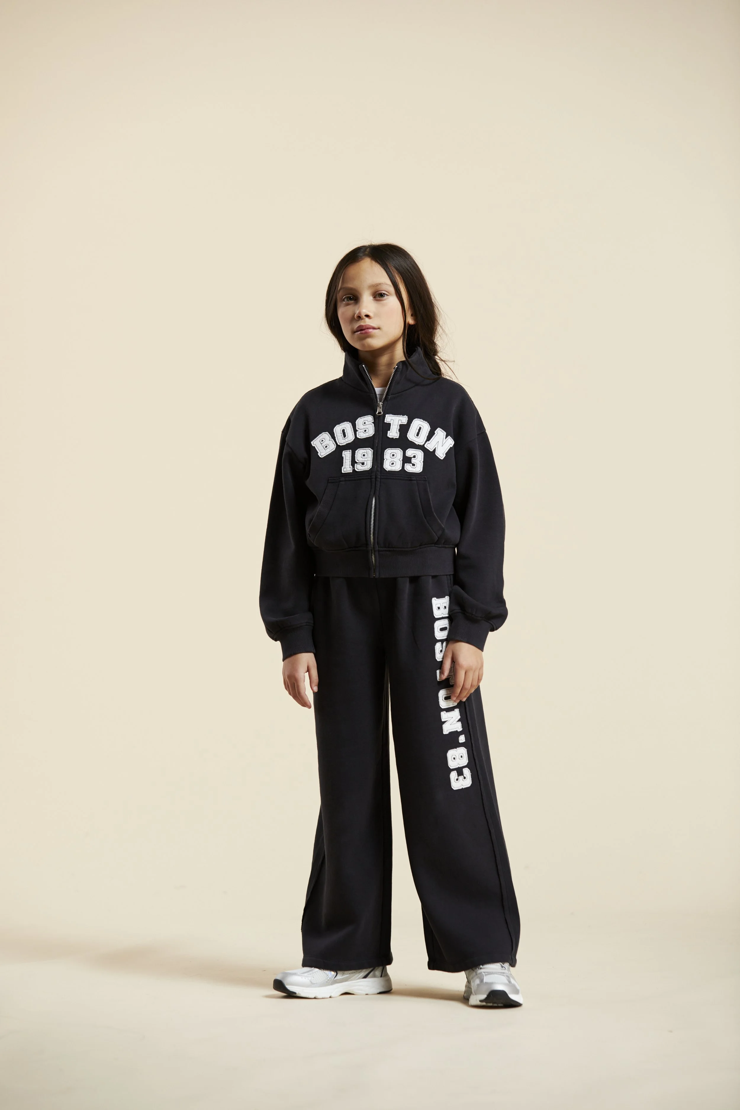 A young girl wearing a black Boston sweatshirt and matching black sweatpants with white lettering, posing against a plain light-colored background. Modelling for leading child model and talent agency SocityUK based in London and Manchester.