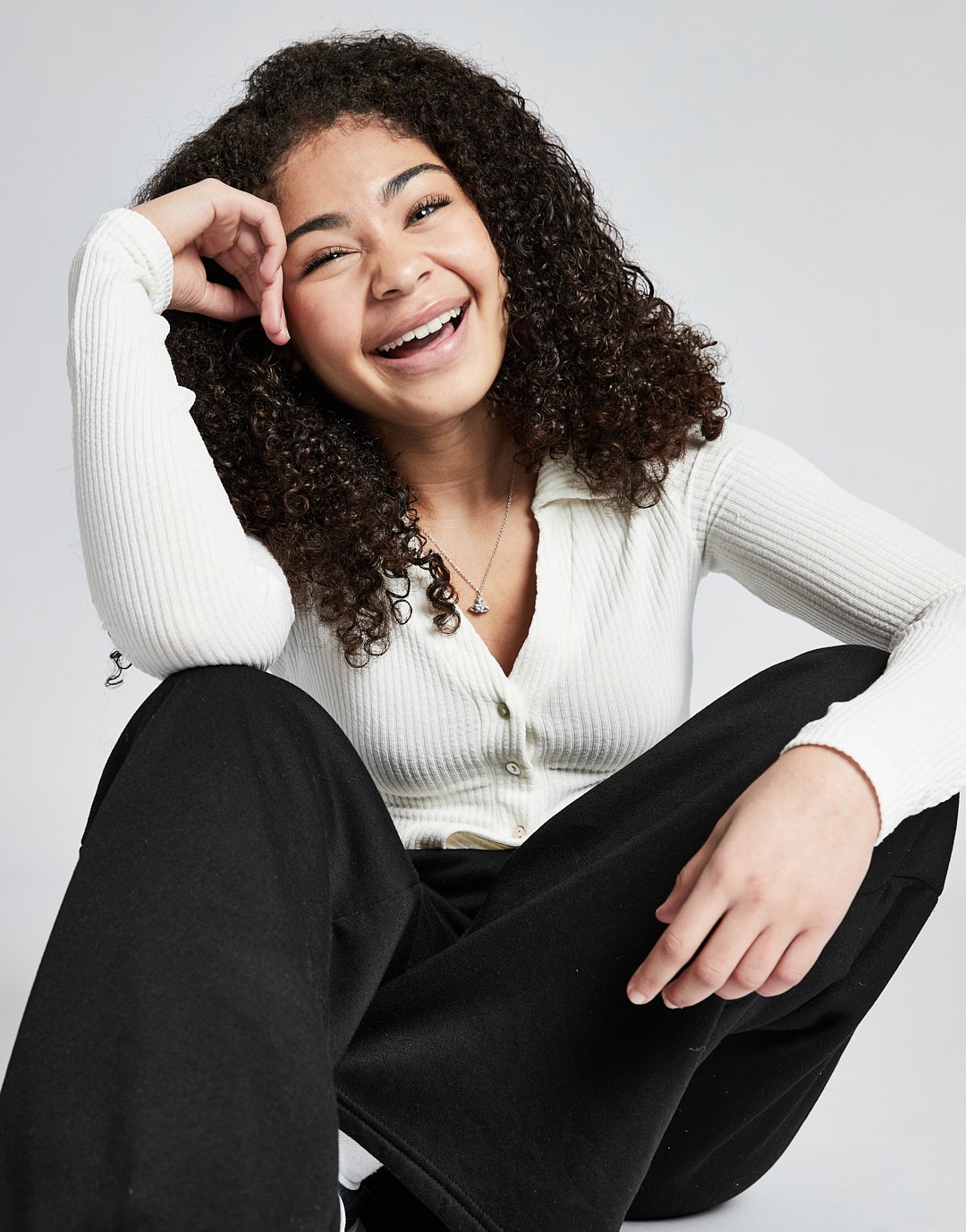 A young woman with curly dark hair, smiling, wearing a white ribbed button-up sweater, black pants, and a necklace, sitting on the floor against a plain gray background.