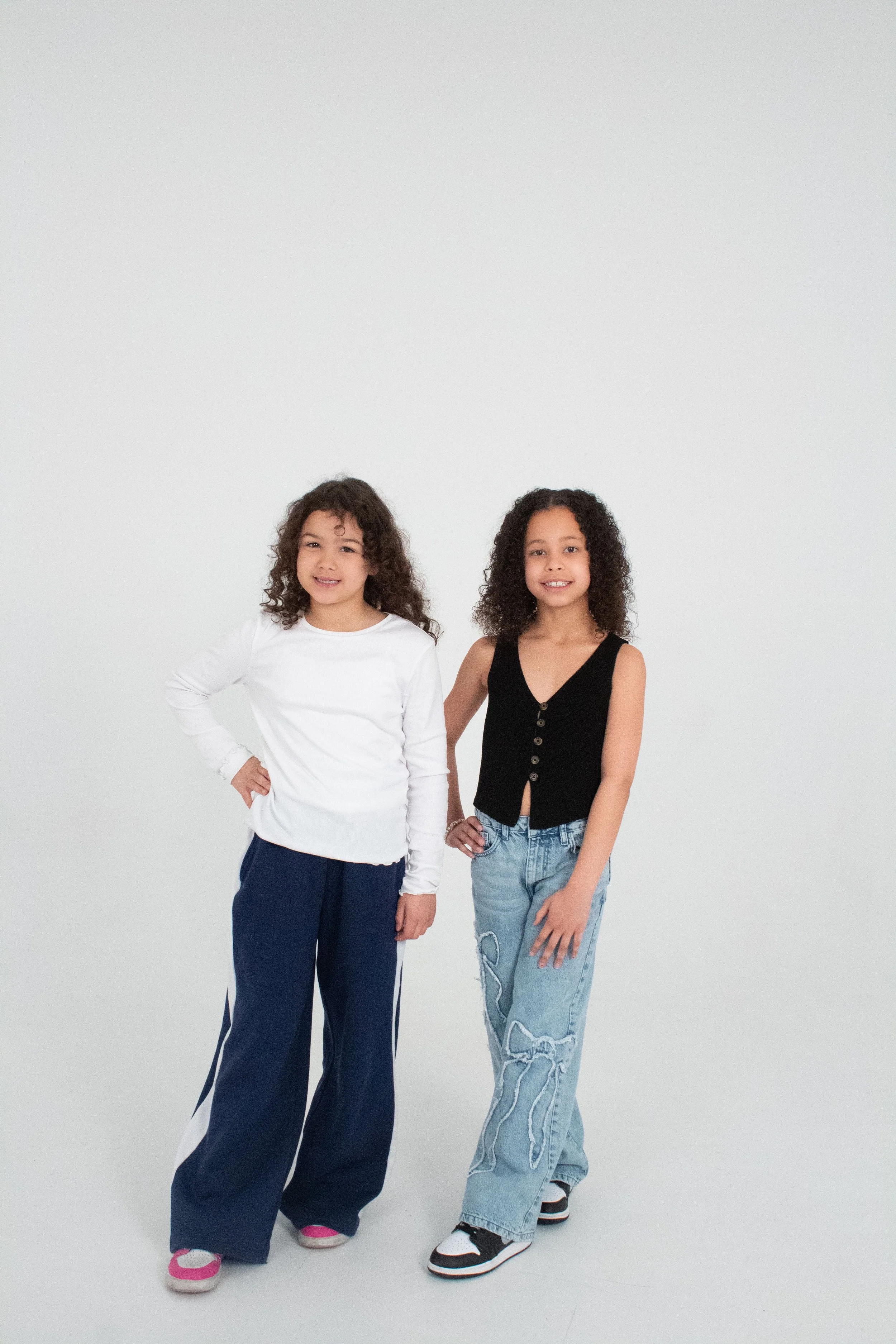 Two young girls with curly hair standing side by side against a plain white background, smiling and wearing casual clothes.