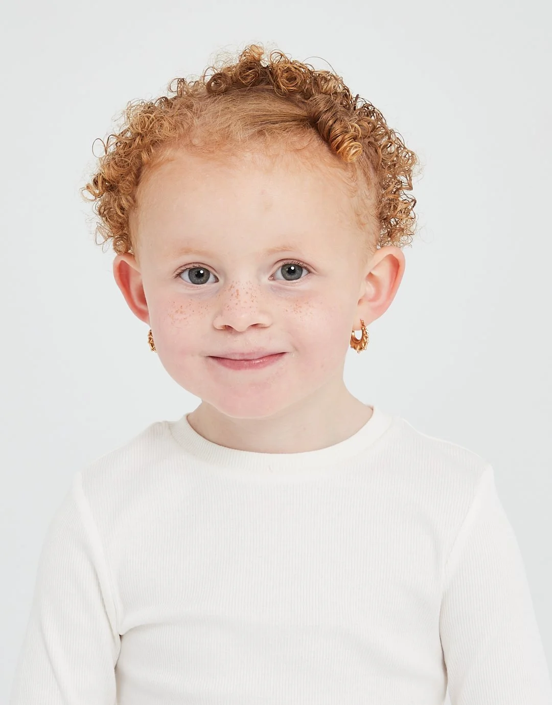 Close-up portrait of a young girl with curly red hair, blue eyes, freckles, gold earrings, and a white long-sleeve shirt, smiling softly against a plain white background. Modelling for leading child model and talent agency SocityUK based in London an