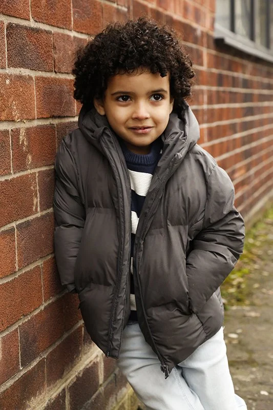 Young boy with curly hair standing against a brick wall, wearing a dark puffer jacket and striped sweater, smiling slightly.