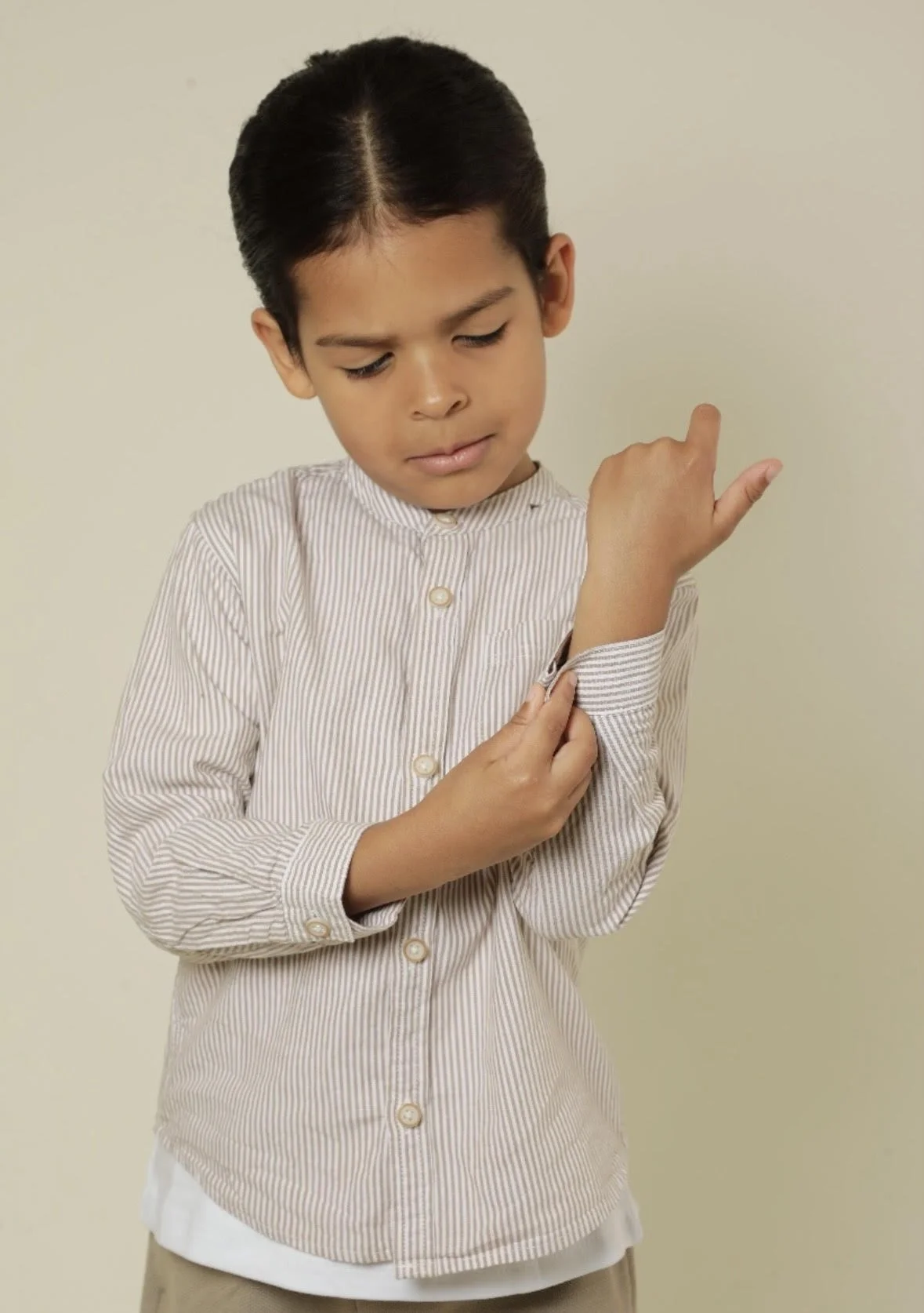 A young boy with dark hair looking down while adjusting the cuff of his striped beige and white shirt.