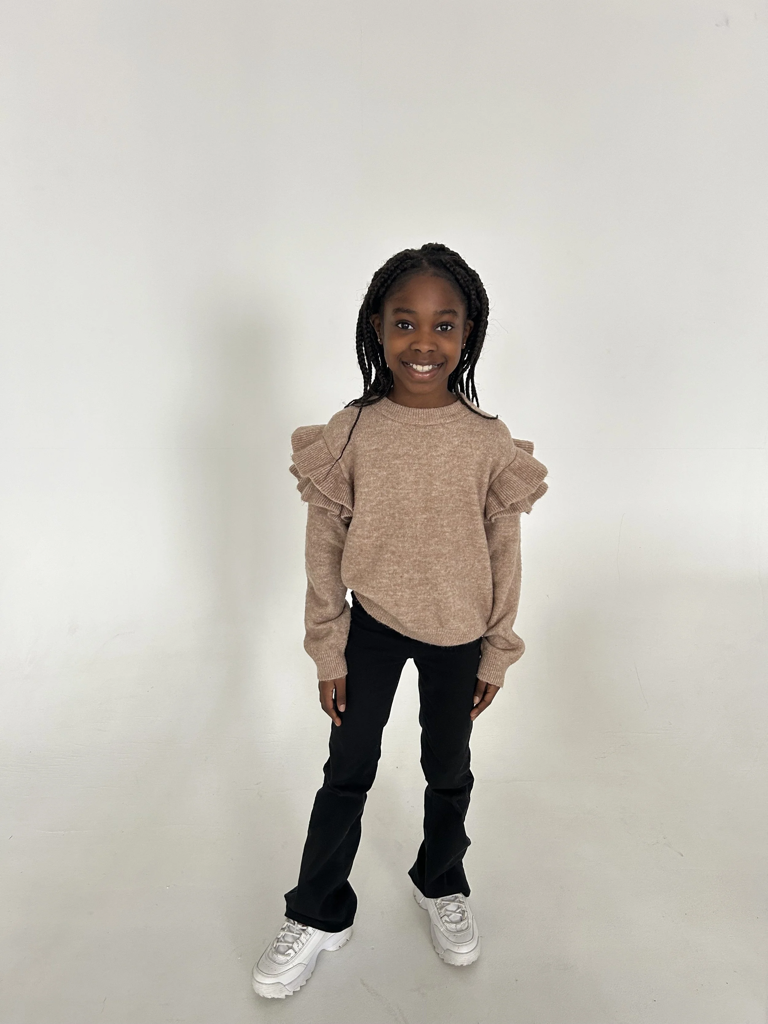 A young girl with dark braided hair smiling, wearing a beige sweater with ruffled shoulders, black pants, and white sneakers, standing against a plain white wall. Model for leading Manchester and London based child model agency Socityuk.