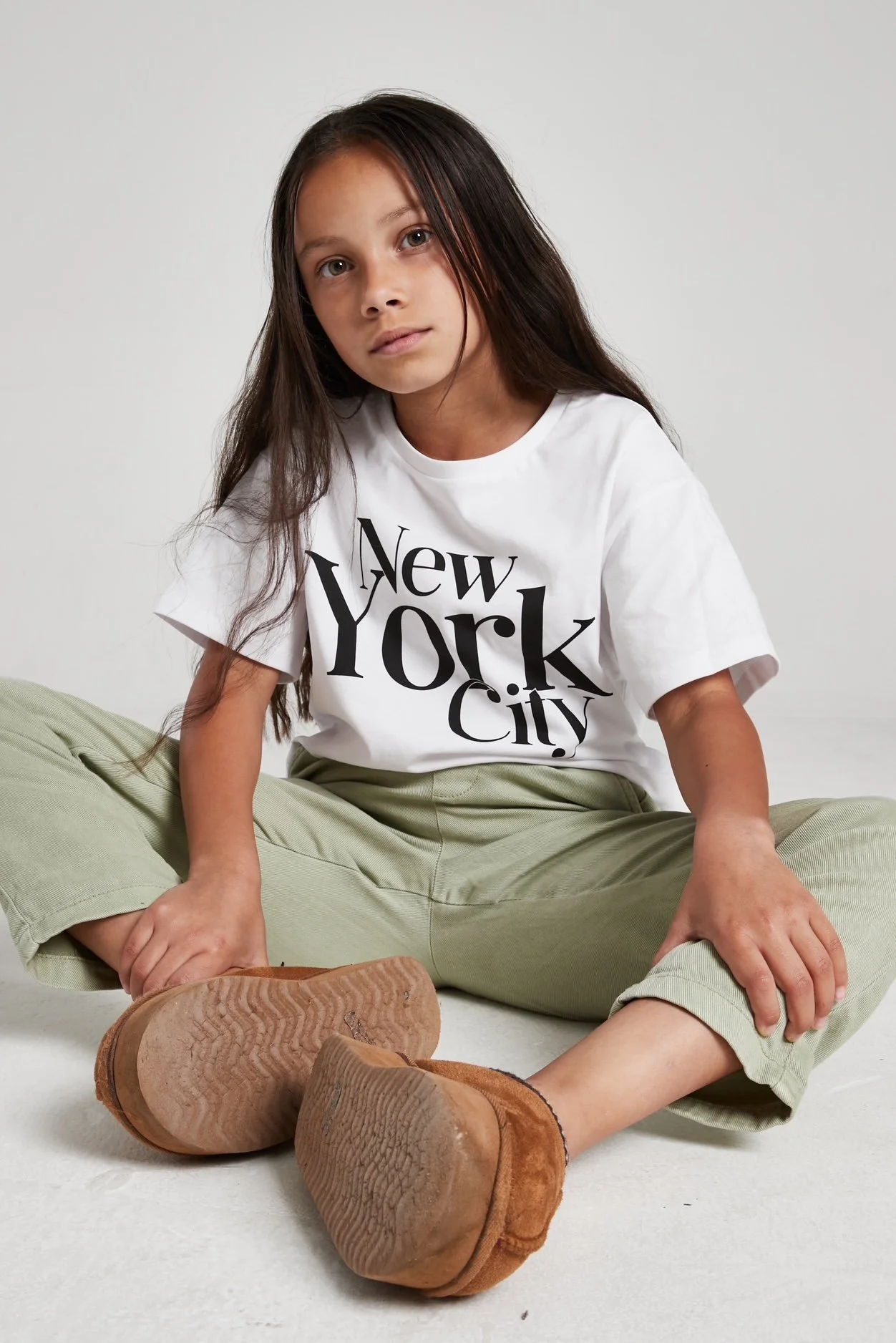 A young girl with long brown hair sitting on the floor, wearing a white t-shirt with 'New York City' printed on it, light green pants, and brown shoes. Modelling for leading child model and talent agency SocityUK based in London and Manchester.