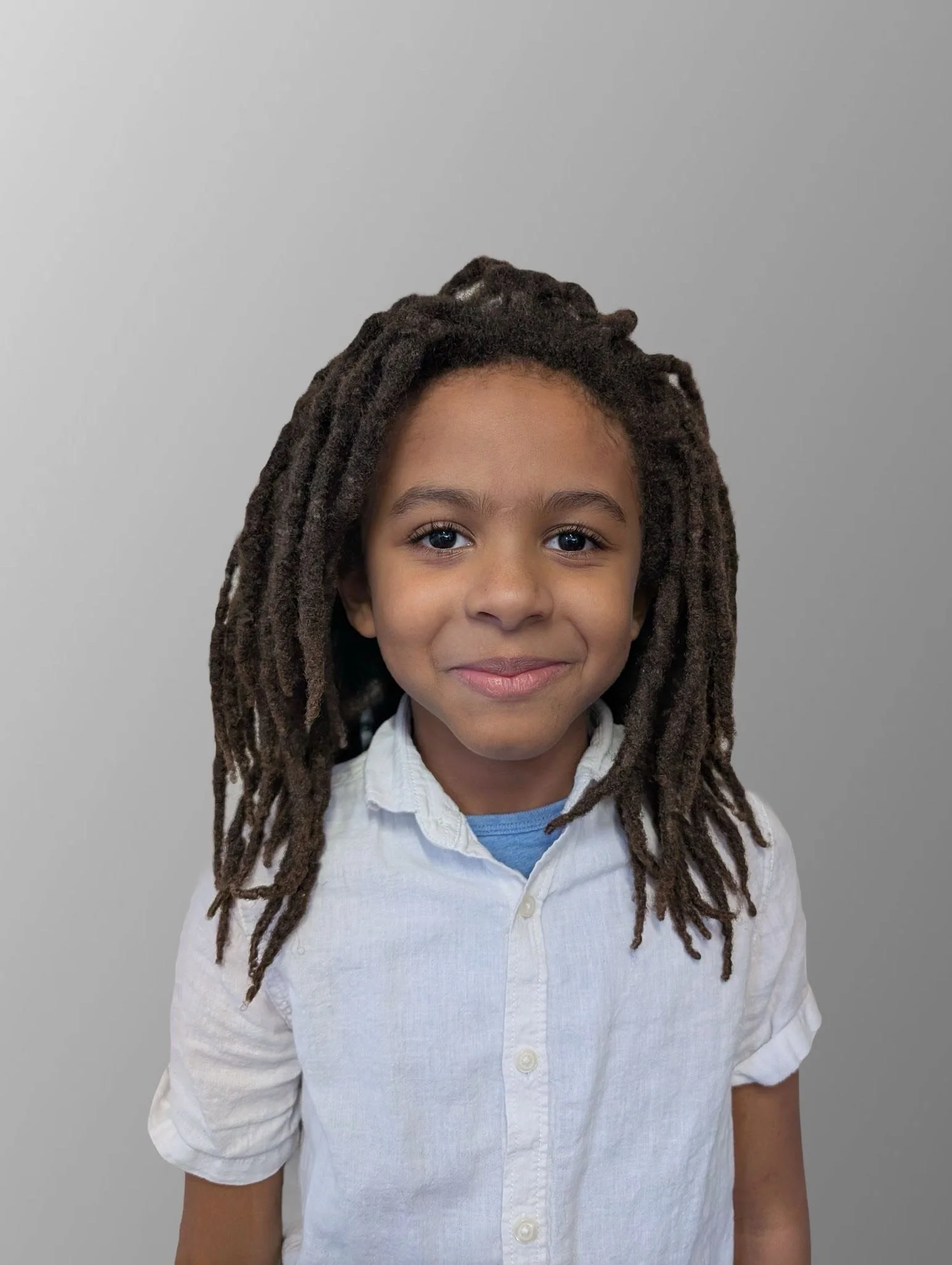 A young boy with dark brown dreadlocks, wearing a light blue shirt and white button-up shirt, smiling against a plain gray background.