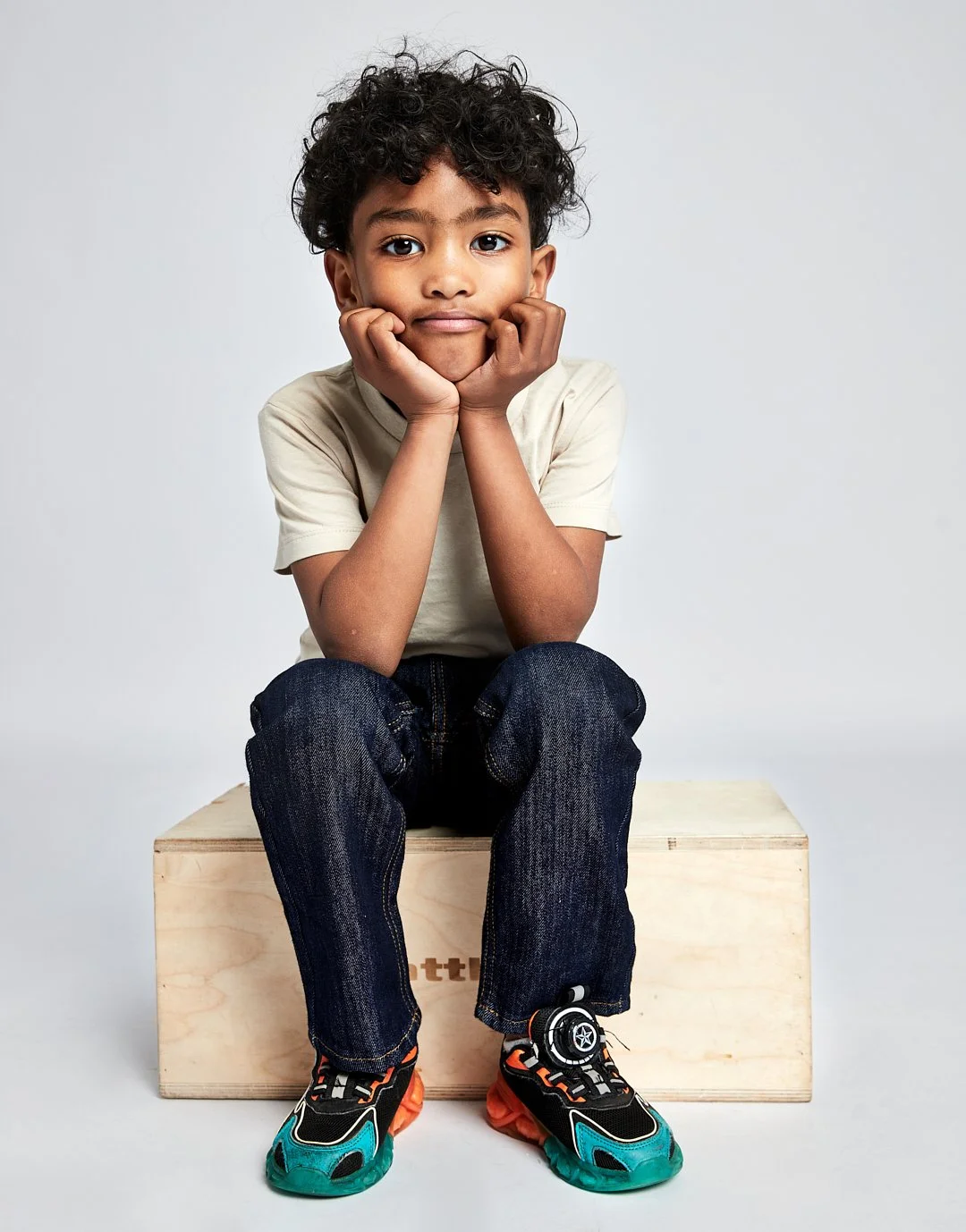 A young boy with curly dark hair sitting on a wooden box against a plain white background, resting his chin on his hands with a thoughtful expression, wearing a cream-colored t-shirt, dark jeans, and colorful sneakers. Modelling for leading child mod