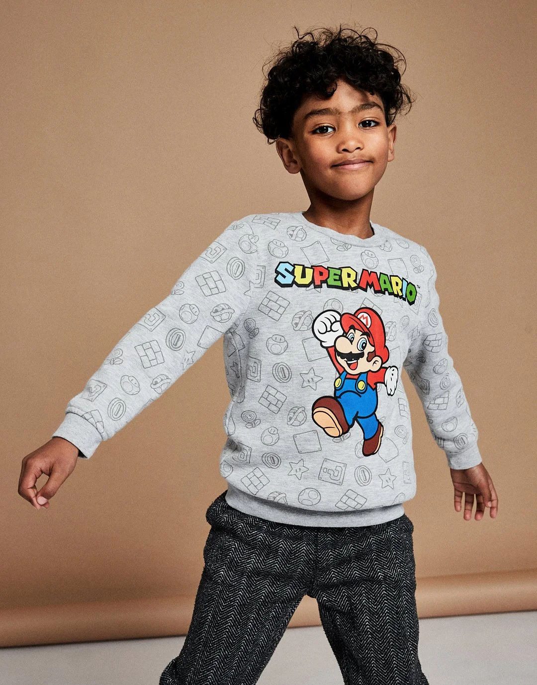 A young boy with curly hair wearing a gray sweatshirt with Super Mario graphics and black and white herringbone pants, standing against a beige background. Modelling for leading child model and talent agency SocityUK based in London and Manchester.