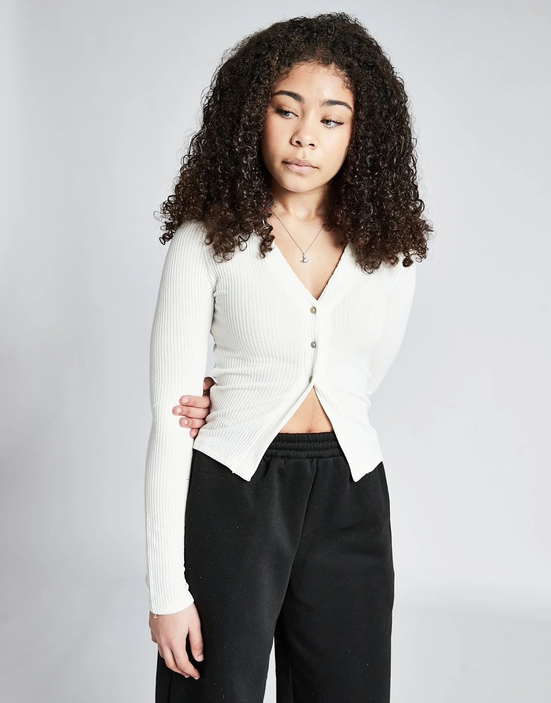 Young woman with curly hair wearing a white button-up top and black pants, standing against a light gray background.