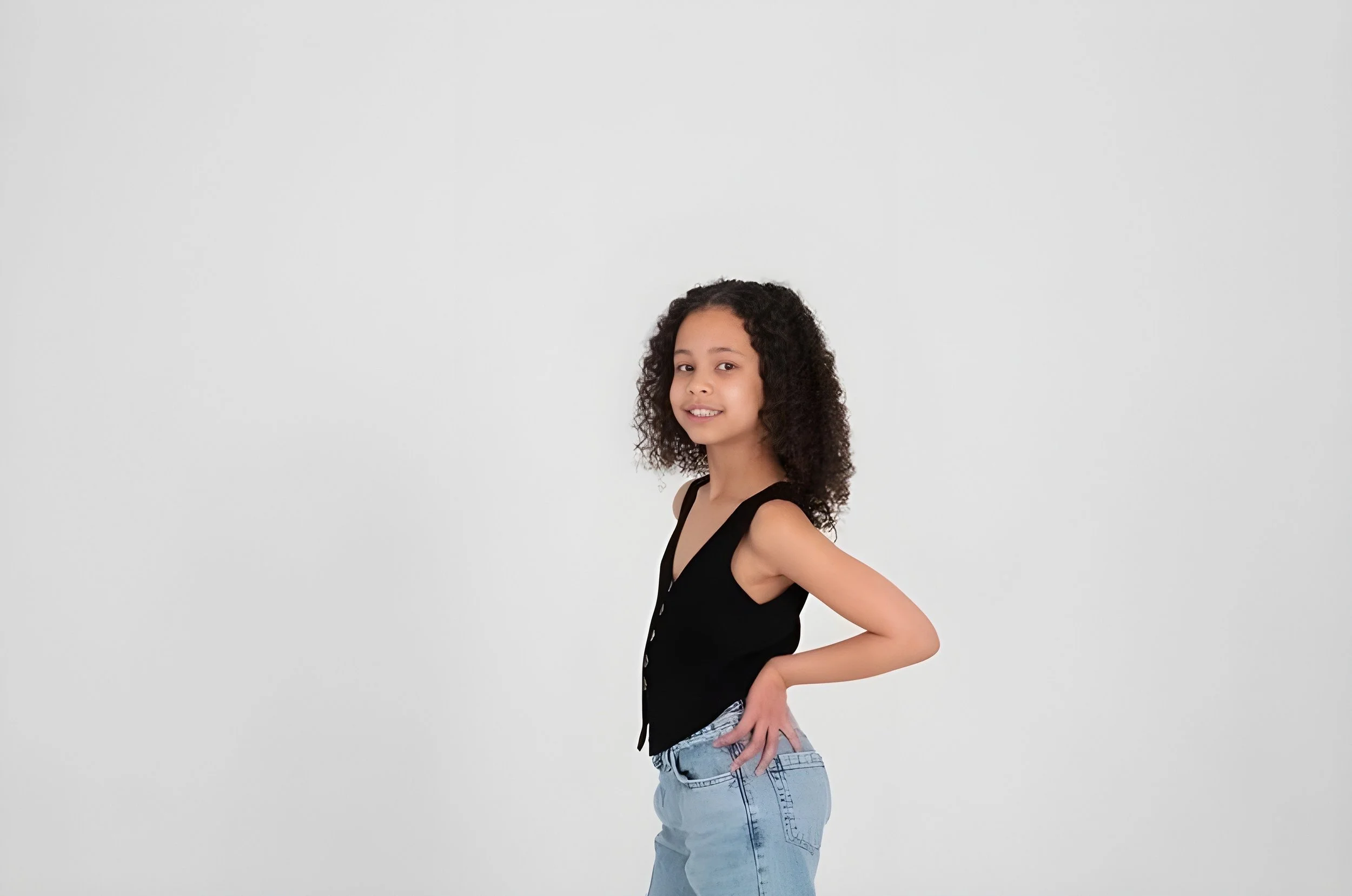 A young girl with curly hair standing against a plain white background, wearing a black sleeveless top and light blue jeans, posing with a slight smile and her right hand on her hip.