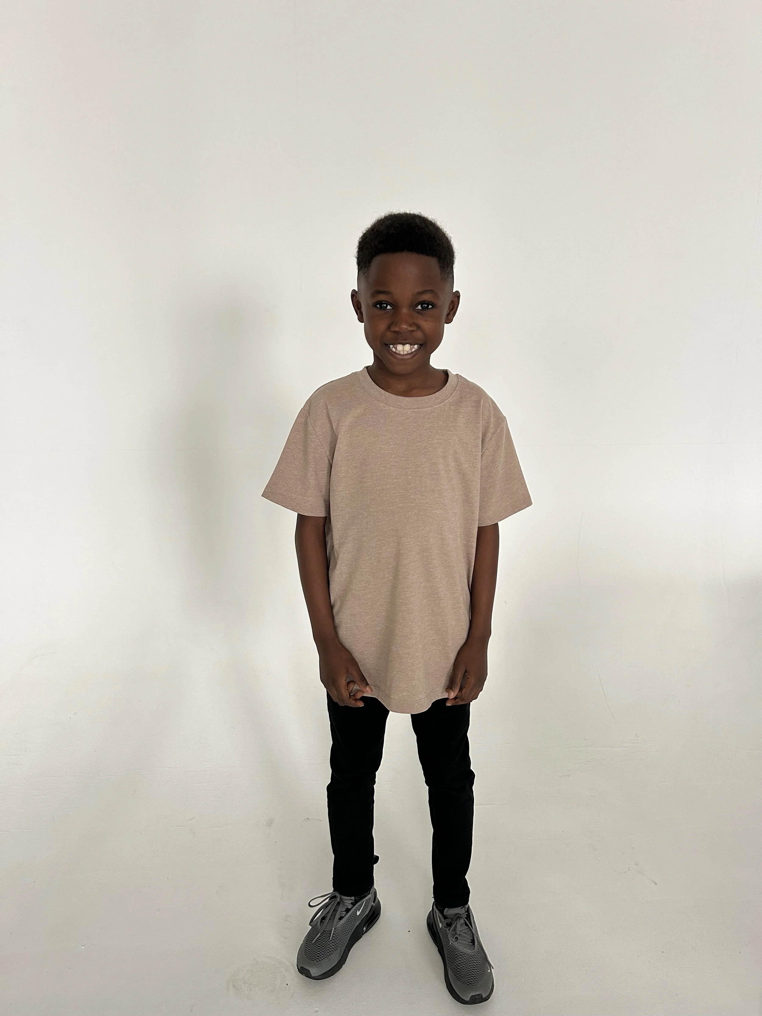 A young boy with dark skin and short hair smiling, wearing a beige T-shirt, black pants, and gray sneakers, standing against a plain white background. Model for leading Manchester and London based child model agency Socityuk.