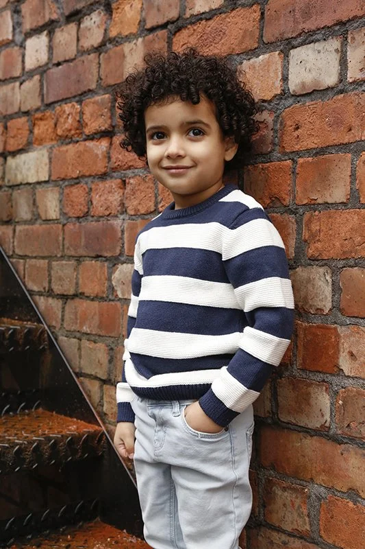 Young boy with curly hair wearing a navy and white striped sweater and light gray pants standing against a red brick wall