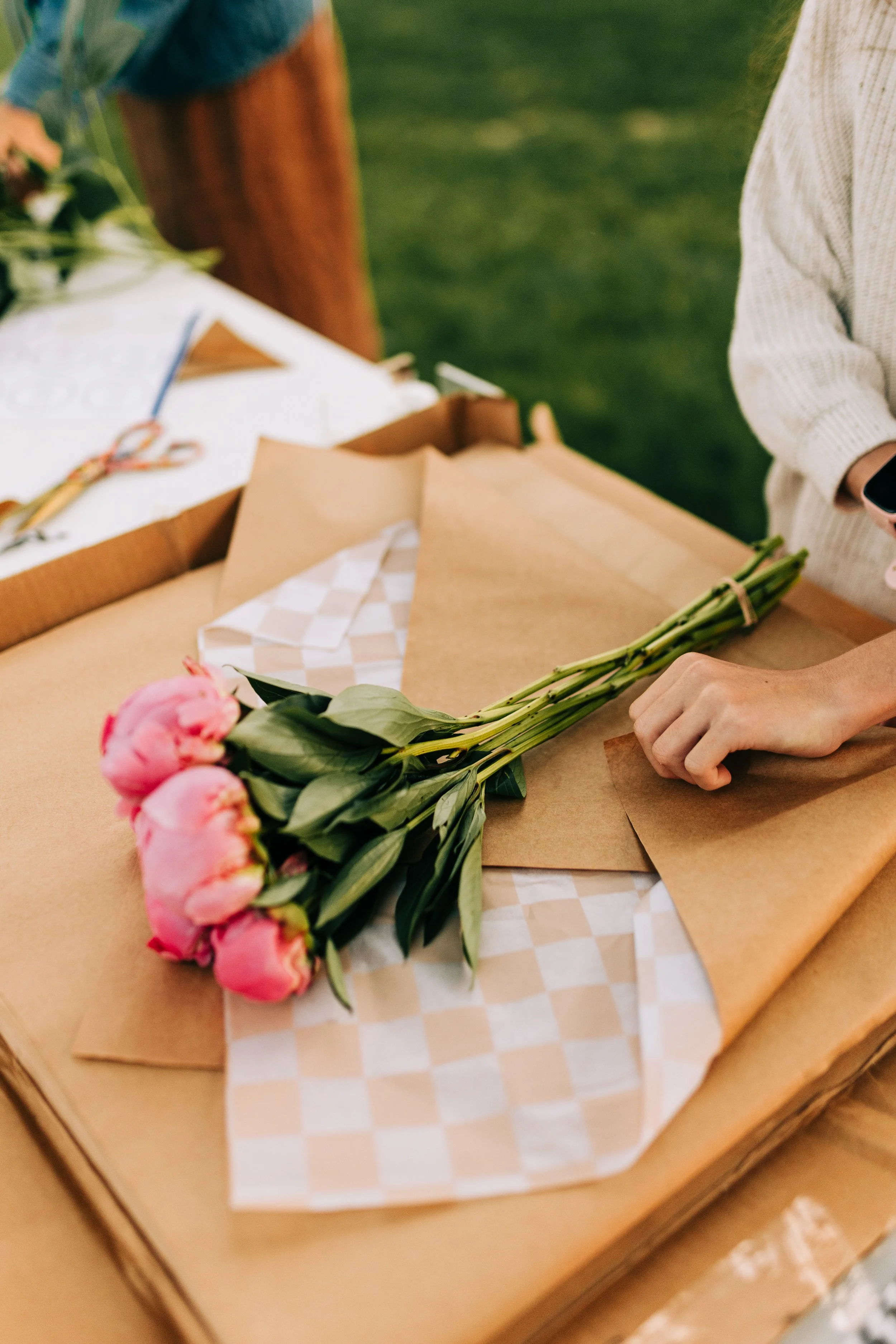 A person wrapping a bouquet of pink peonies on a table with brown paper and checked tissue paper, in an outdoor setting.