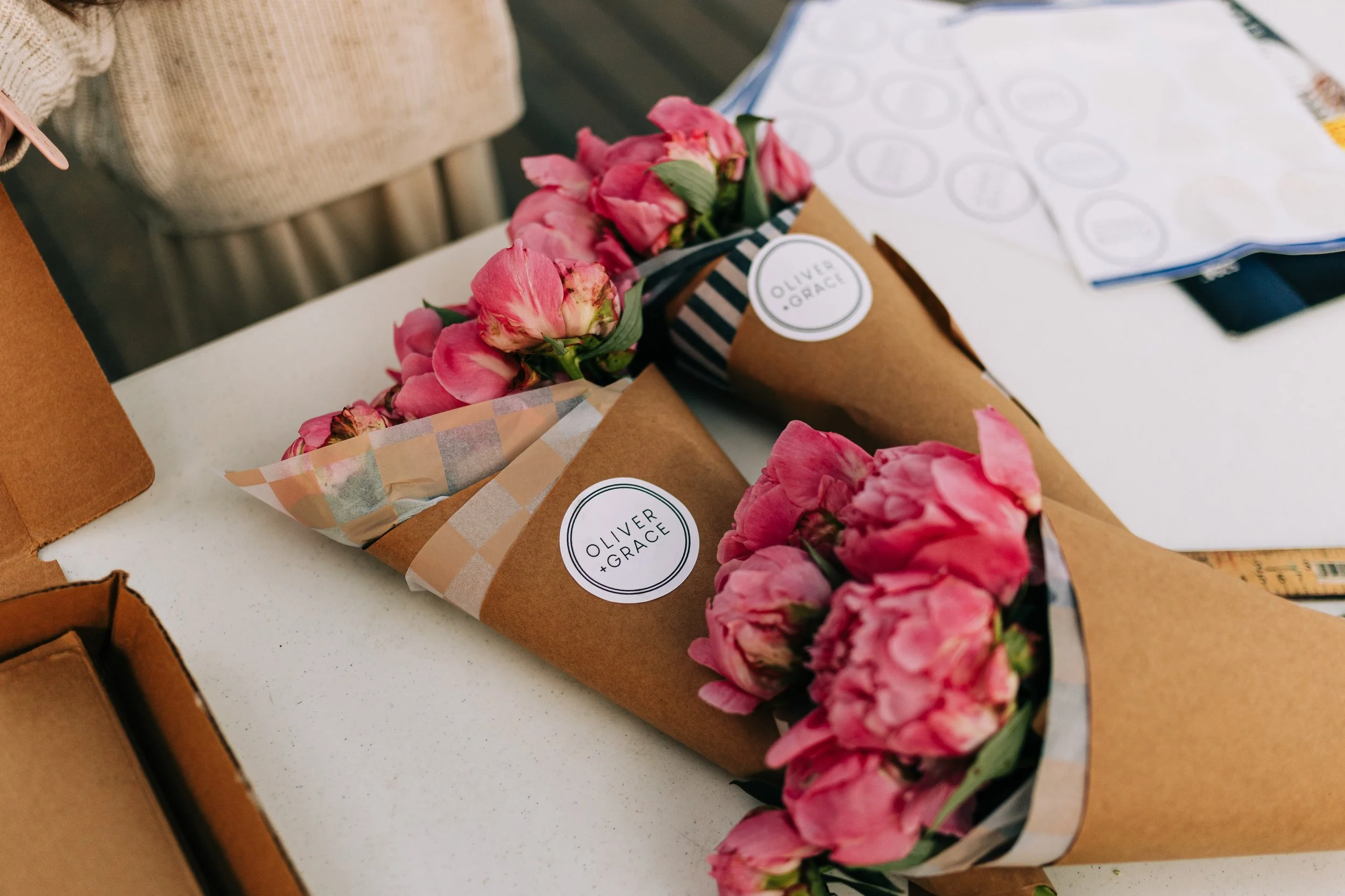 Two bouquets of pink peonies wrapped in brown paper with a sticker that says 'OLIVER + GRACE' on a white table. There are some boxes and papers nearby.