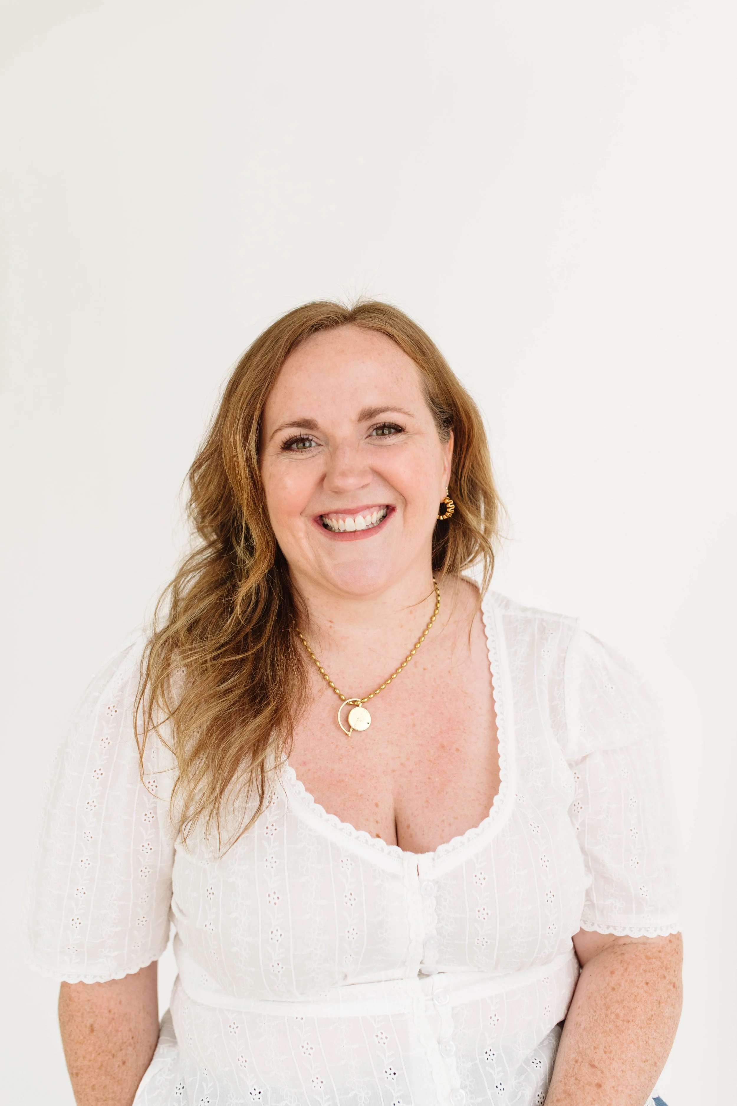 A woman with long, wavy, reddish-brown hair smiling at the camera, wearing a white embroidered blouse, gold jewelry, and sitting against a plain white background.