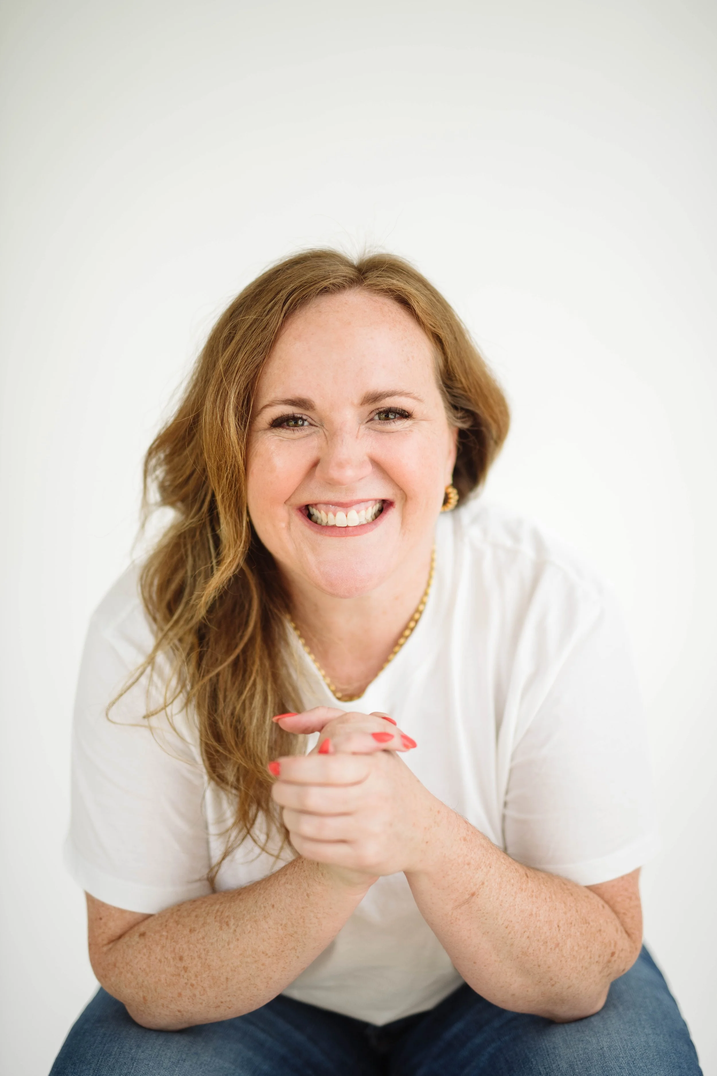 Close-up of a smiling woman with red hair, wearing a white top, gold necklace, and gold earrings, sitting against a white background.