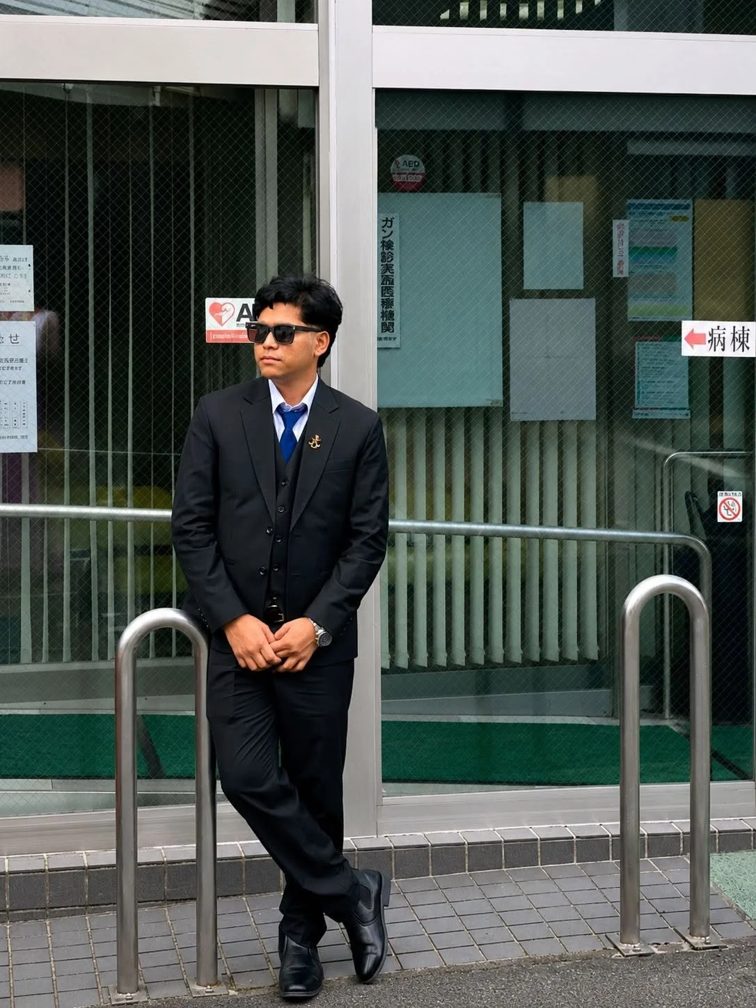 Man in black suit and sunglasses standing outside a glass building, leaning on bike racks.