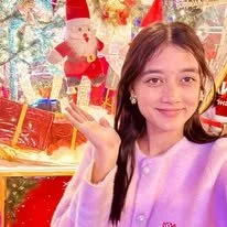 A girl with long dark hair smiling and making a peace sign with her hand, in front of a festive Christmas display with Santa Claus and wrapped presents.