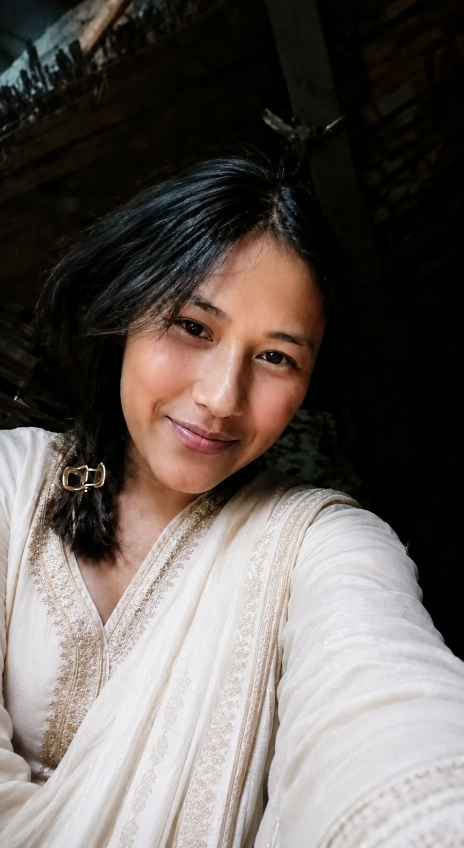 A woman with short black hair smiling at the camera, wearing a cream-colored embroidered traditional outfit, with a dark wooden background.