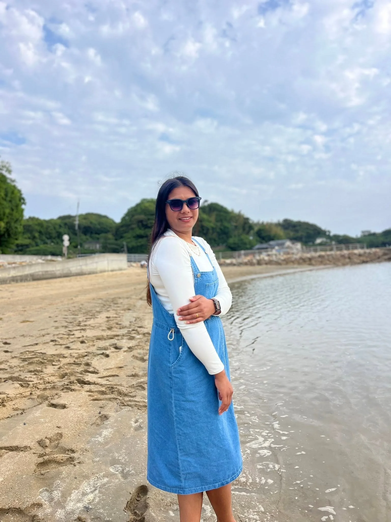 A woman standing on a sandy beach near the water, wearing sunglasses, a white long-sleeved shirt, and a blue denim overall dress, with her arms crossed, smiling at the camera under a partly cloudy sky.