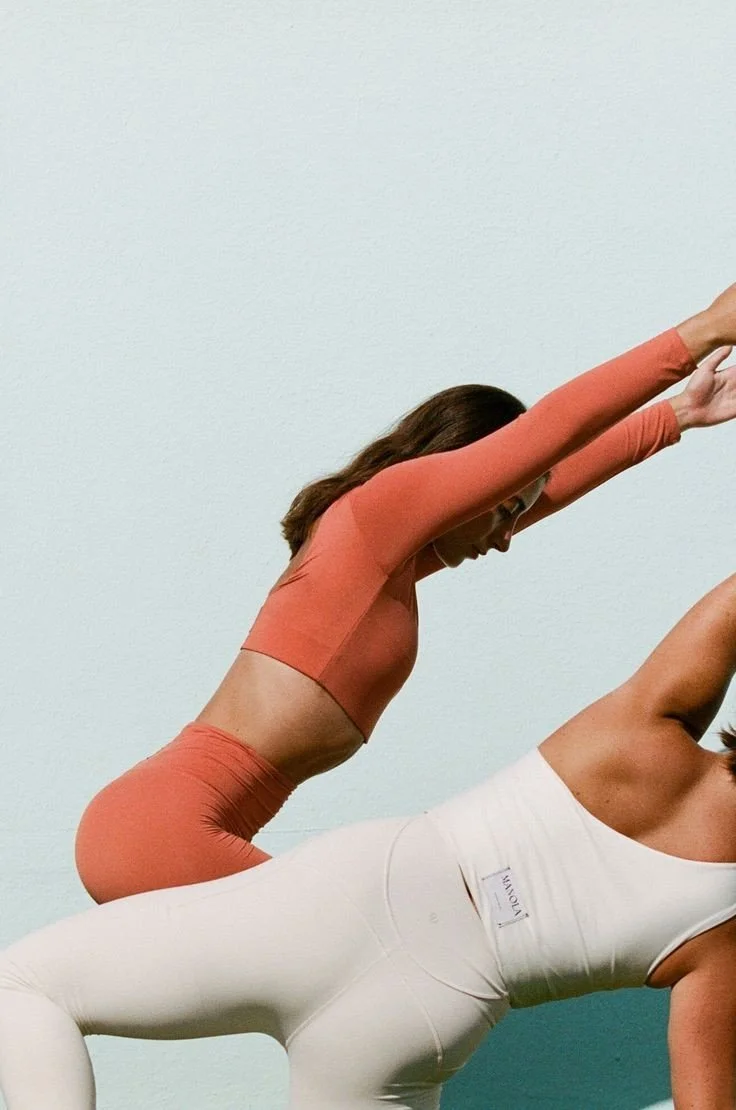 Two women practicing yoga against a plain wall, one in a plank pose and the other in a downward dog pose.