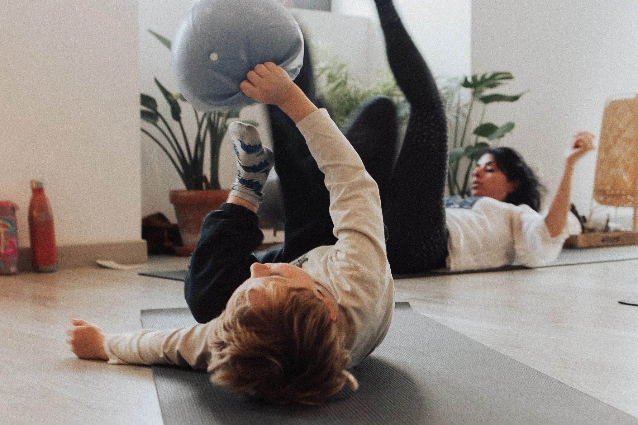 Two children playing on yoga mats indoors, one lying on her back and holding a ball, the other lying on her side with arms raised, with houseplants and a lamp in the background.