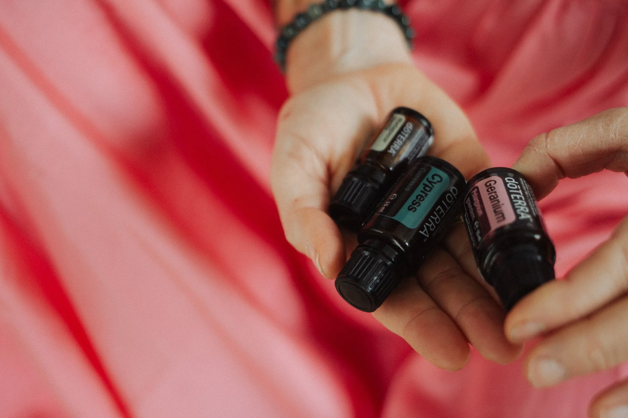 A person holding three small bottles of essential oils labeled Gua Sha, Geranium, and another oil, with a pink fabric background.
