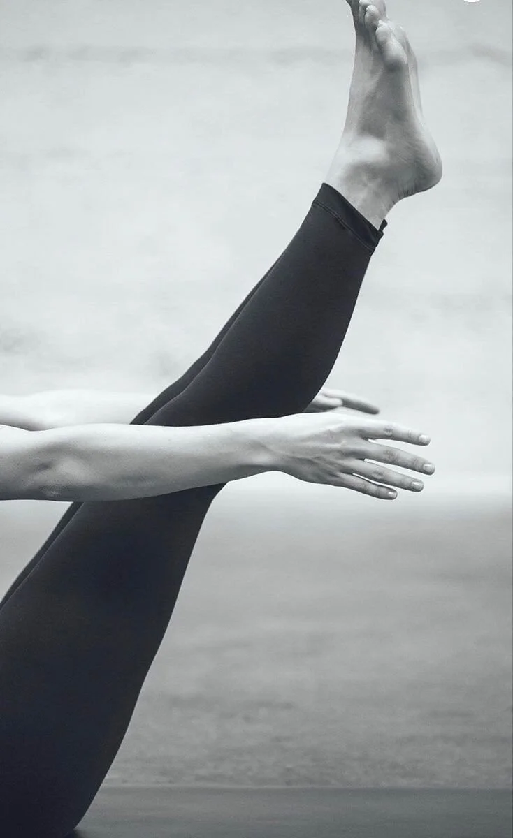 A black and white photo of a person doing a yoga pose, with one leg extended vertically and the other supporting their body, against a blurred outdoor background.