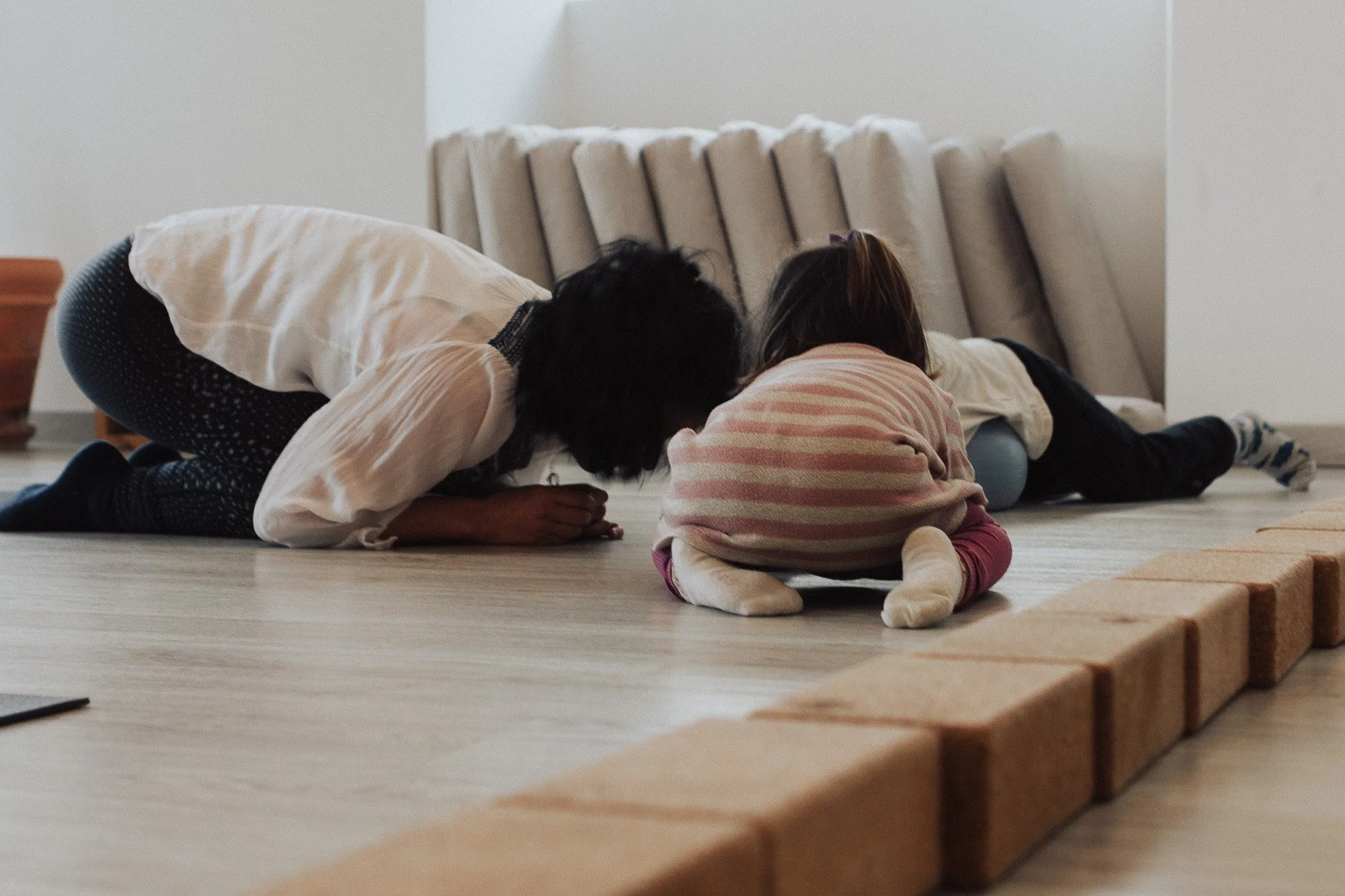 Three children kneeling on the floor with their foreheads touching, appearing to be praying or engaging in a quiet activity together in a room with a bed and blankets in the background.