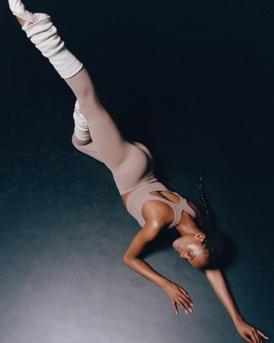 A woman in a taupe athletic outfit with braids lying on the floor, stretching with one arm extended and her eyes closed.