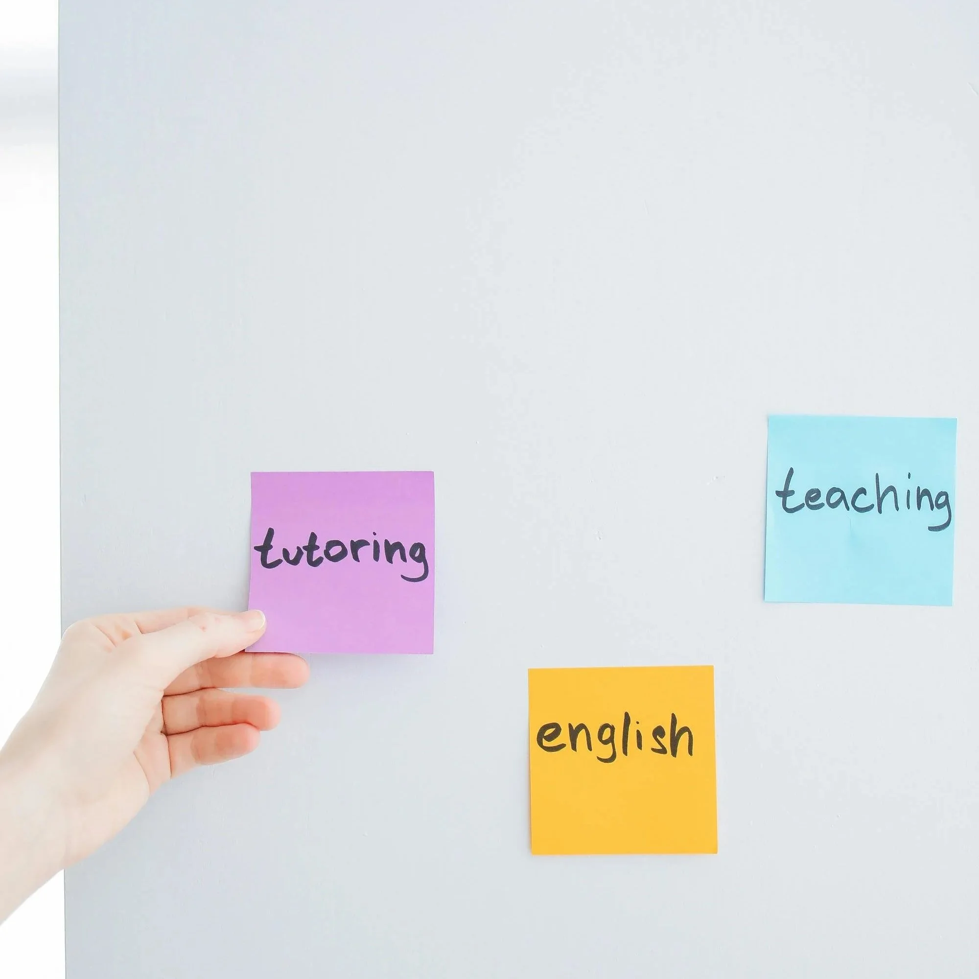 Ein violetter Haftnotizzettel mit der Aufschrift „Nachhilfe“ wird von Hand auf eine weiße Tafel geklebt, zusammen mit weiteren blauen und gelben Haftnotizzetteln mit den Aufschriften „Unterricht“ und „Englisch“.