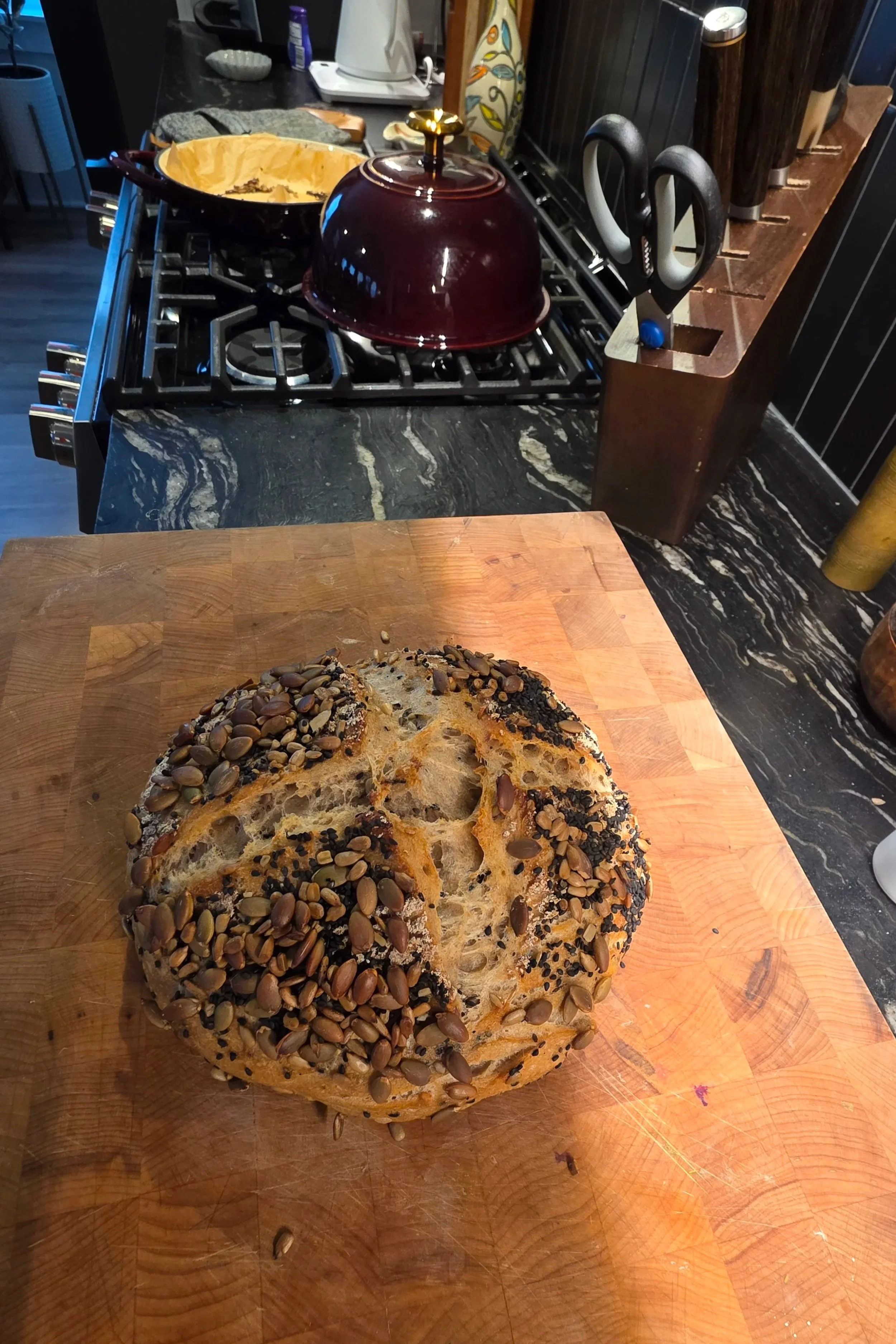 Seeded sourdough bread on wooden cutting board with Dutch oven in home kitchen