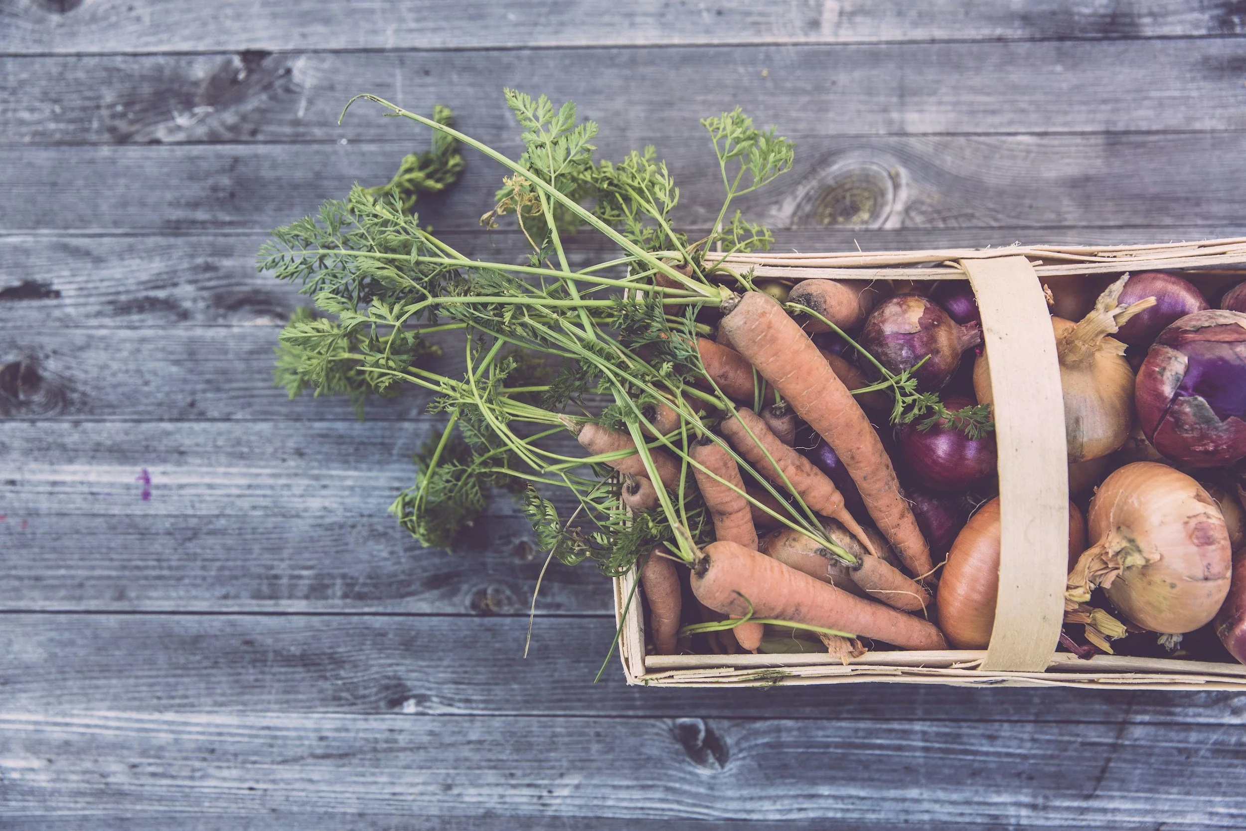A basket of freshly harvested carrots, onions, and shallots on a wooden surface.
