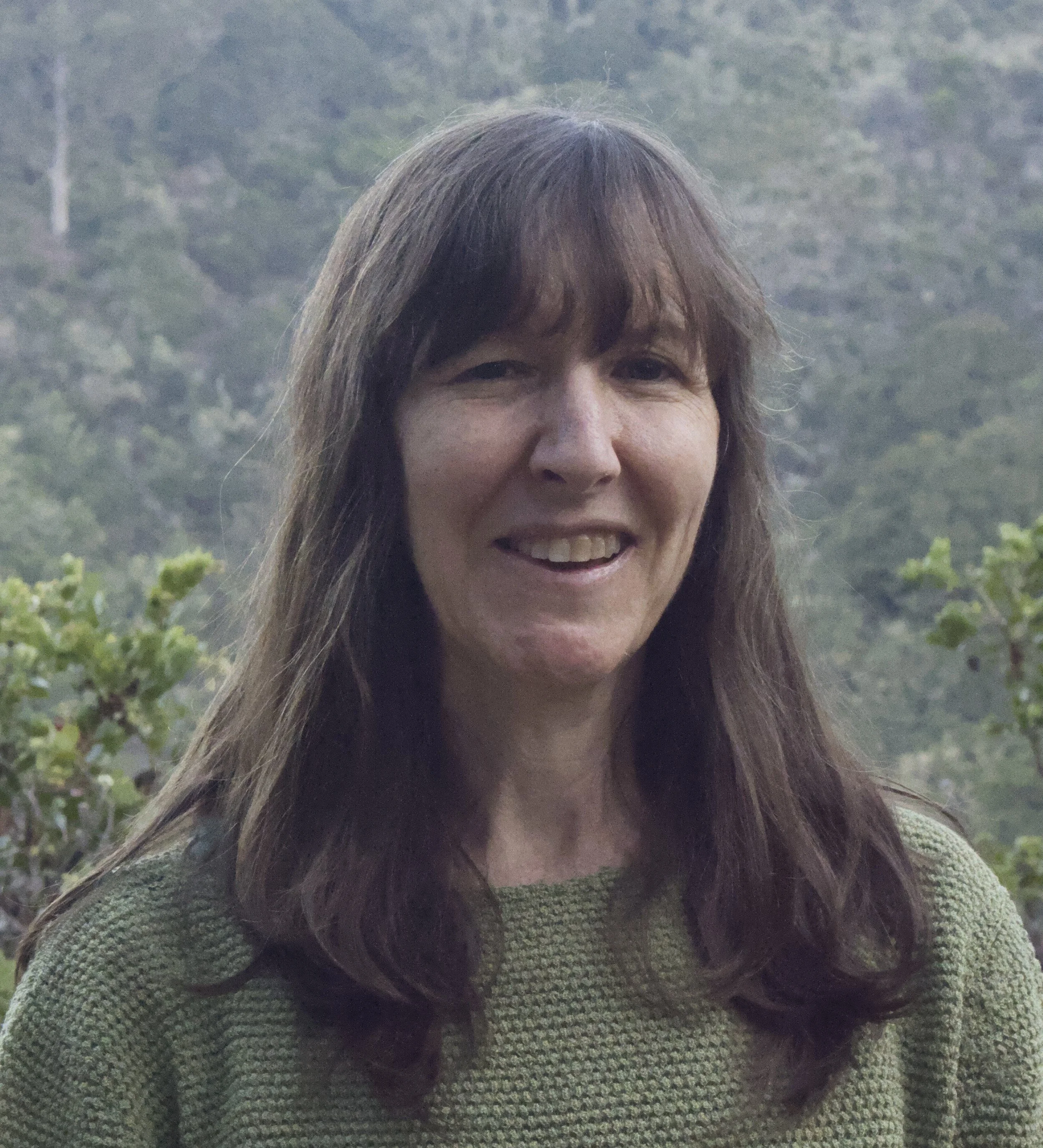 Close-up of a woman with short brown hair and a small smile, wearing a purple top and a silver earring with a turquoise stone, standing in front of a bookshelf filled with various books.