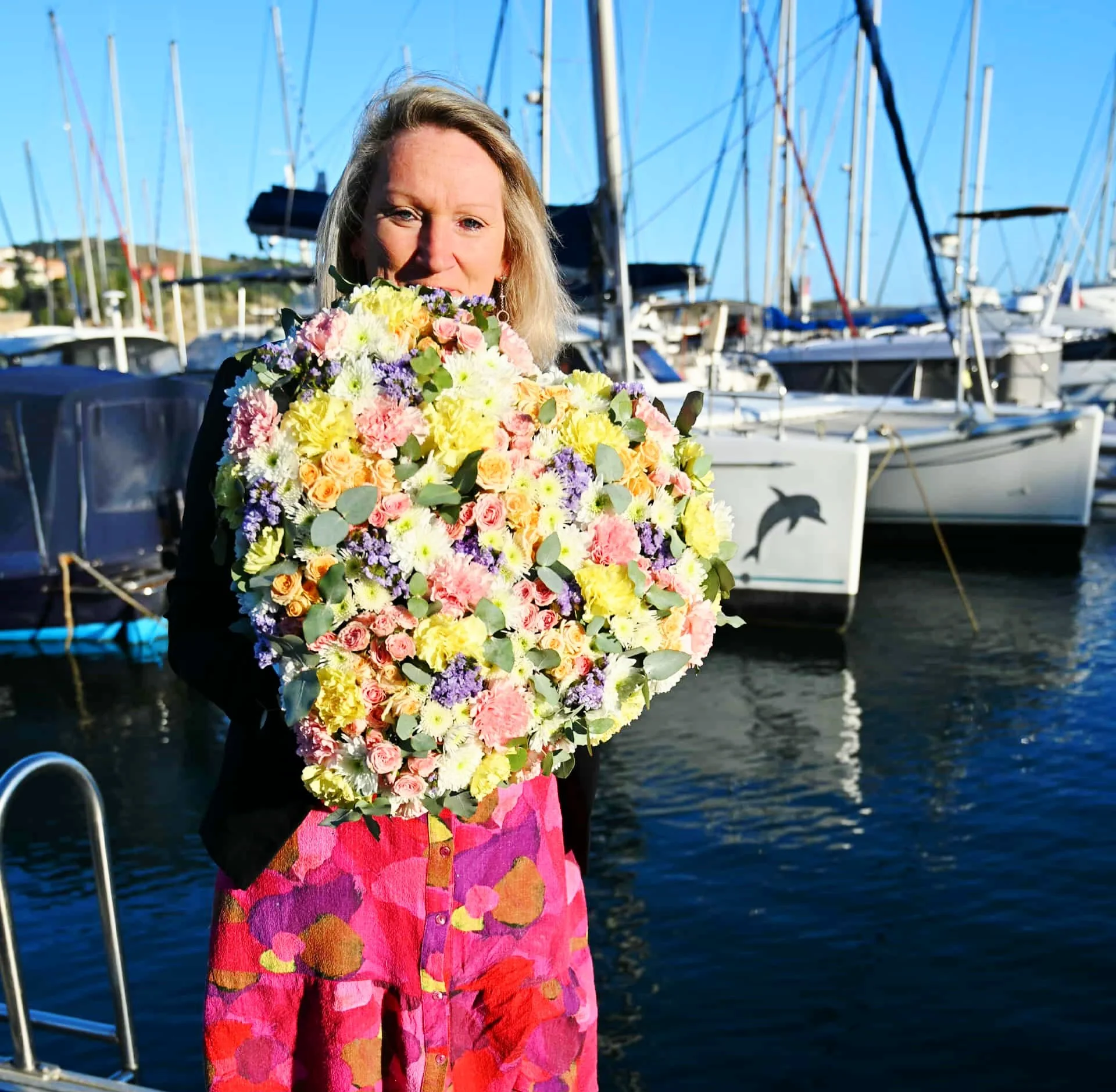 Séverine, fleuriste à Banyuls-sur-Mer, tenant une création en forme de cœur aux tons pastel sur le port de plaisance