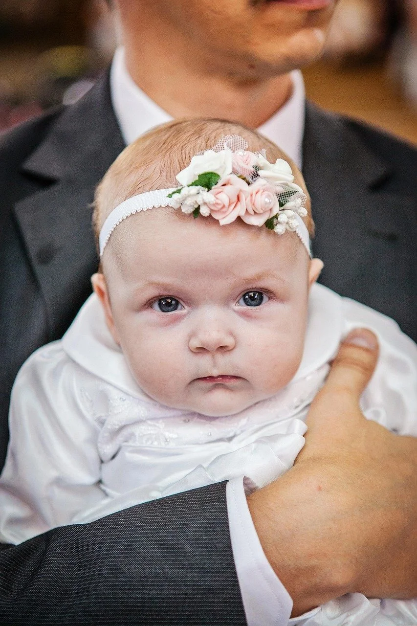 Bébé avec un bandeau floral blanc porté sur la tête, tenant dans les bras un adulte en costume noir lors d'une cérémonie, probablement un baptême ou une mariage..Réalisé par Violette et Abricot Fleuriste bouquetière à Banyuls-sur-Mer