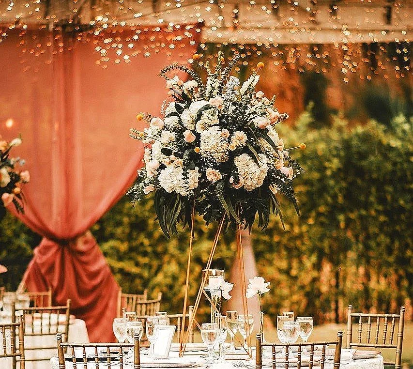 Décoration de mariage avec un grand centre de table floral sur une table dressée, sous un ciel lumineux avec des guirlandes lumineuses.