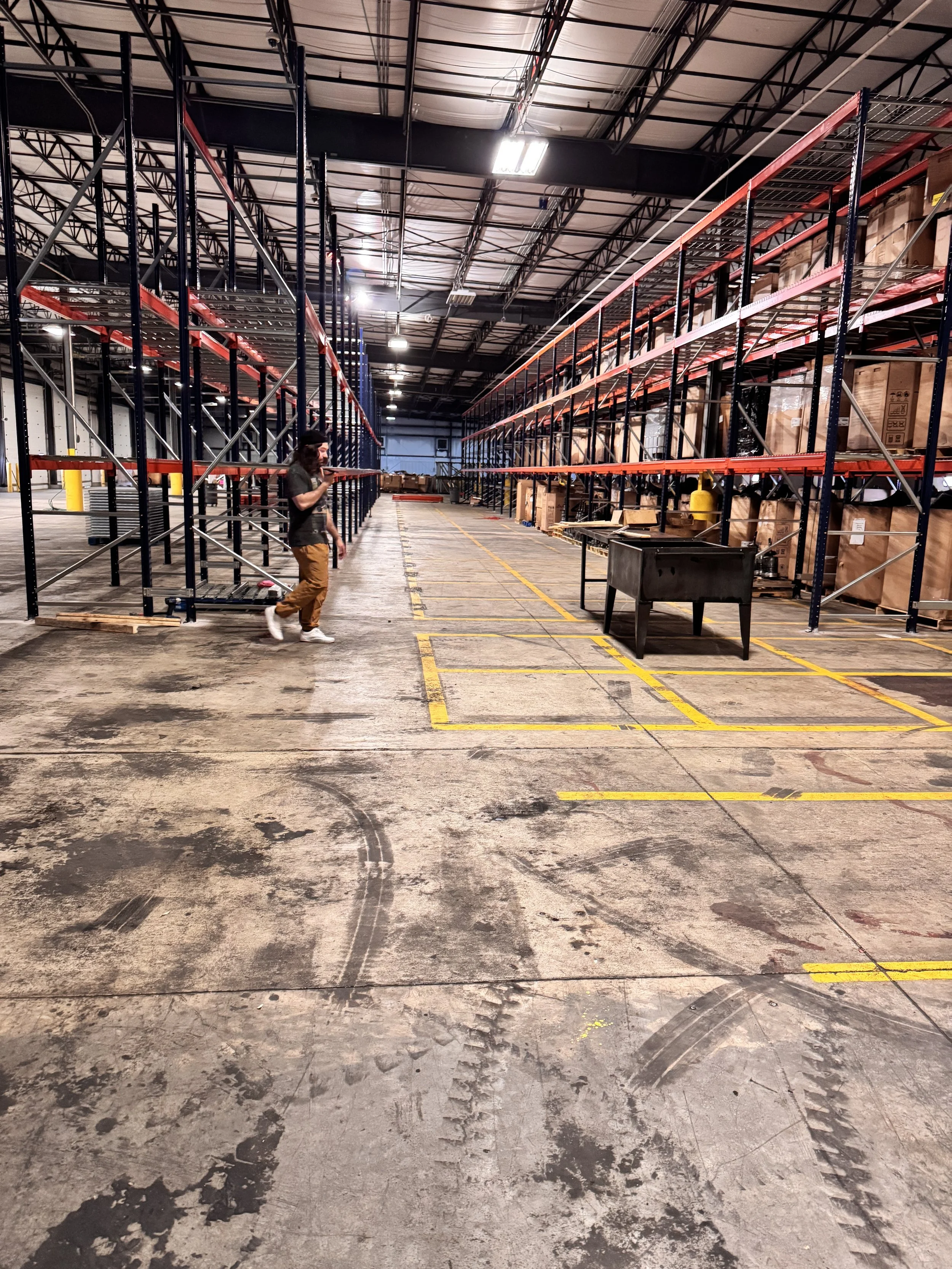 Empty warehouse with tall metal shelving units, a worker walking through, a black work table, and yellow marked bays on the floor.