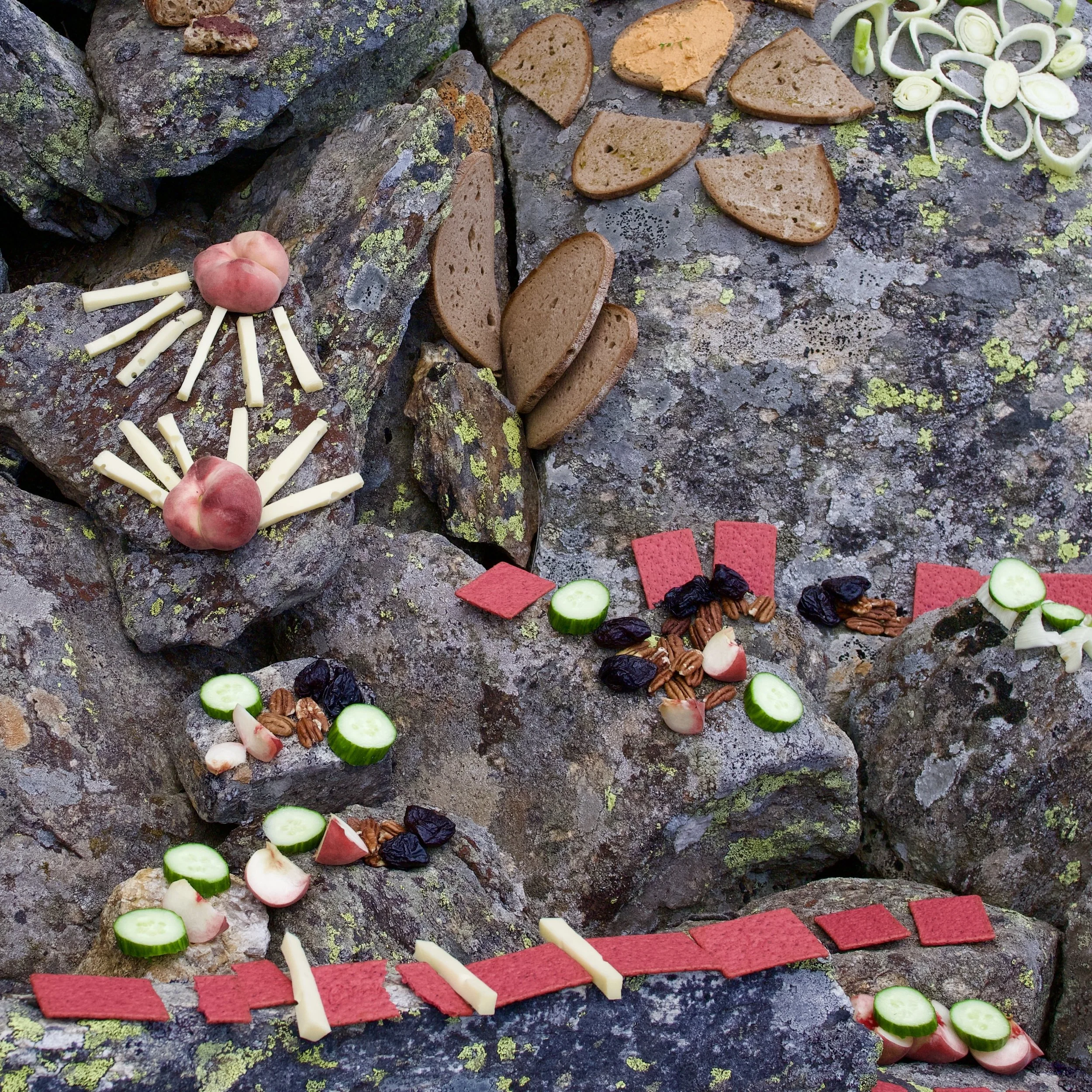 Kunstvolle Anordnung von Lebensmitteln auf Steinen, darunter Radieschen, Käse, Gurkenscheiben, getrocknete Pflaumen, Nüsse und Pappkartons in verschiedenen Farben.